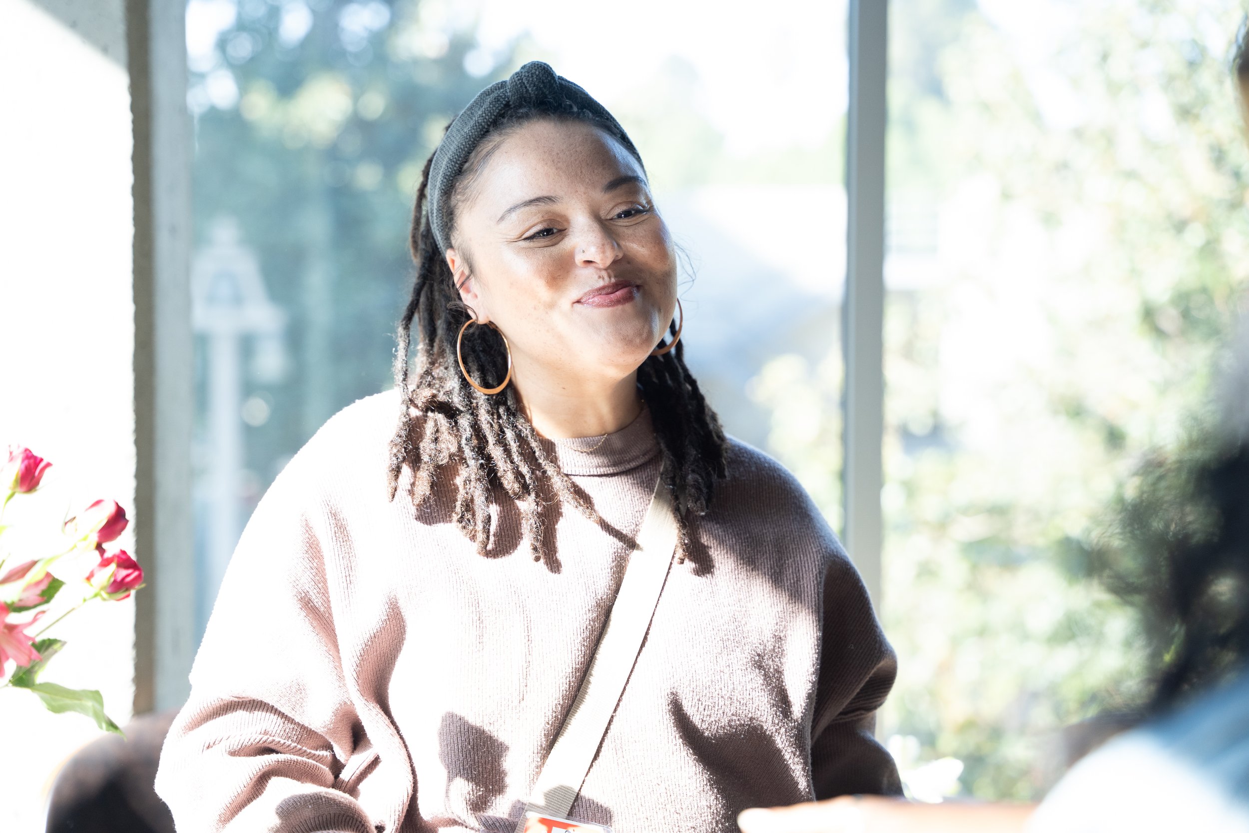 A woman with dreadlocks, wearing a gray headband, gold hoop earrings, and a beige sweater, smiling inside a brightly lit room near a window with trees outside.