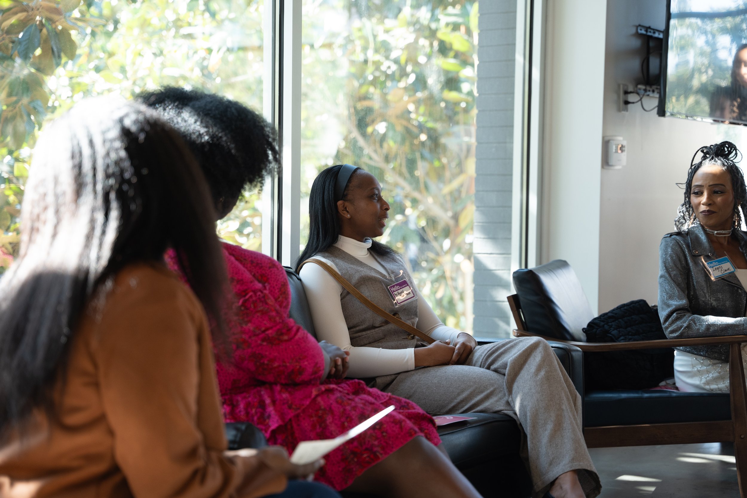 Four women sitting on a black leather couch and chairs in a brightly lit room with large windows and greenery outside, engaged in conversation.