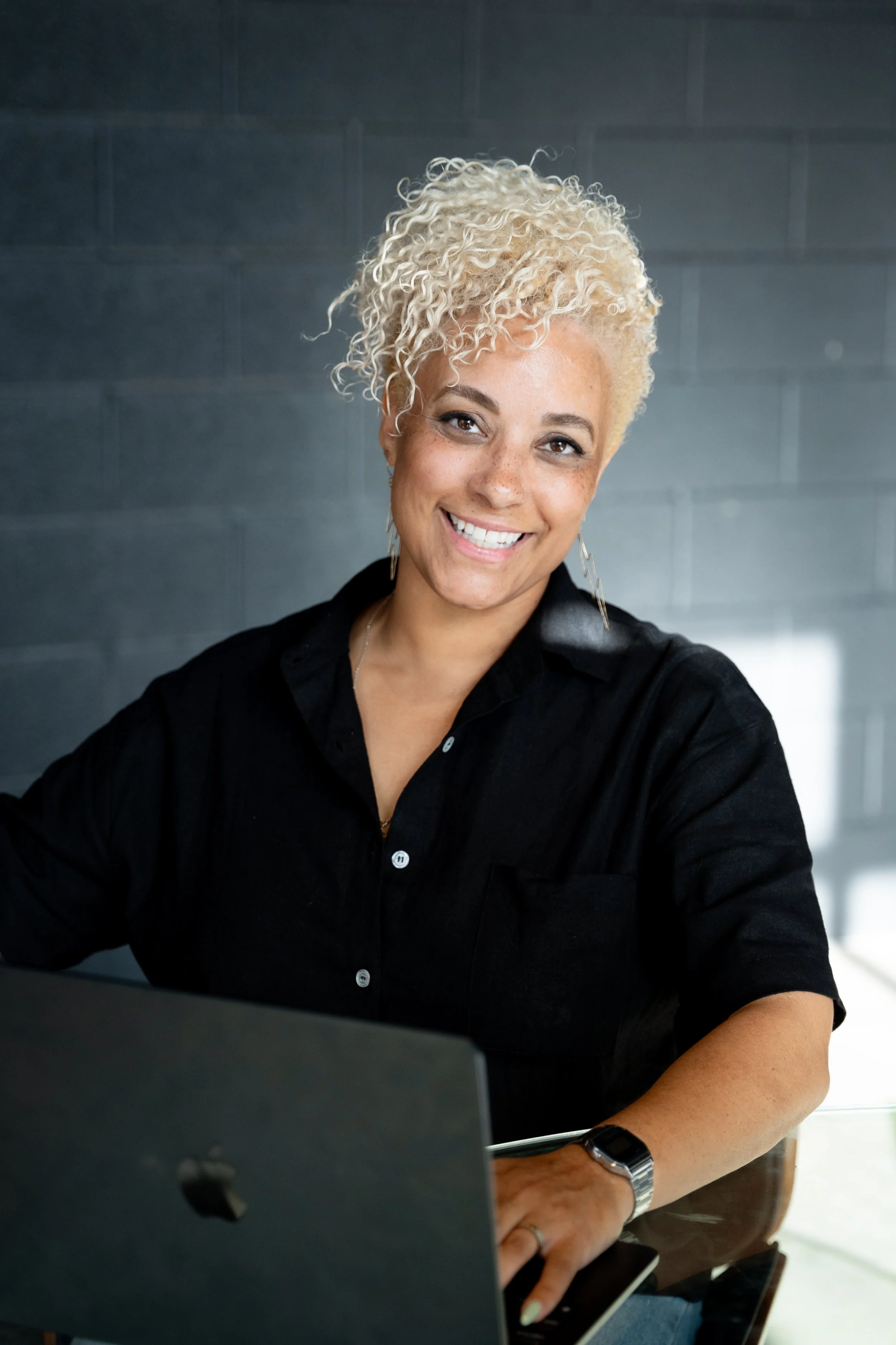 A woman with short, curly blonde hair smiling at the camera, sitting at a desk with a laptop.