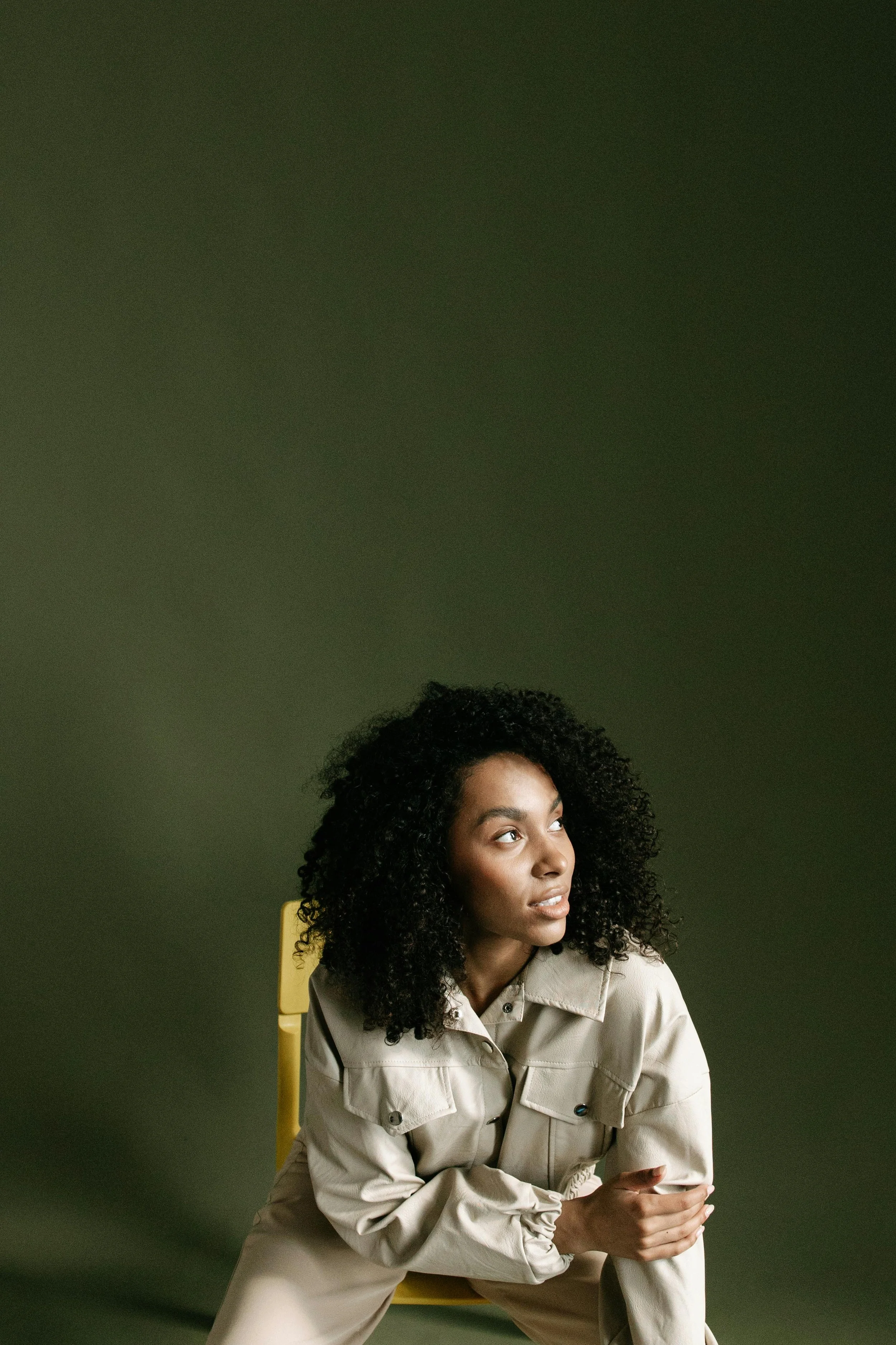 A woman with curly black hair sitting on a yellow chair against a green background, looking to her left.