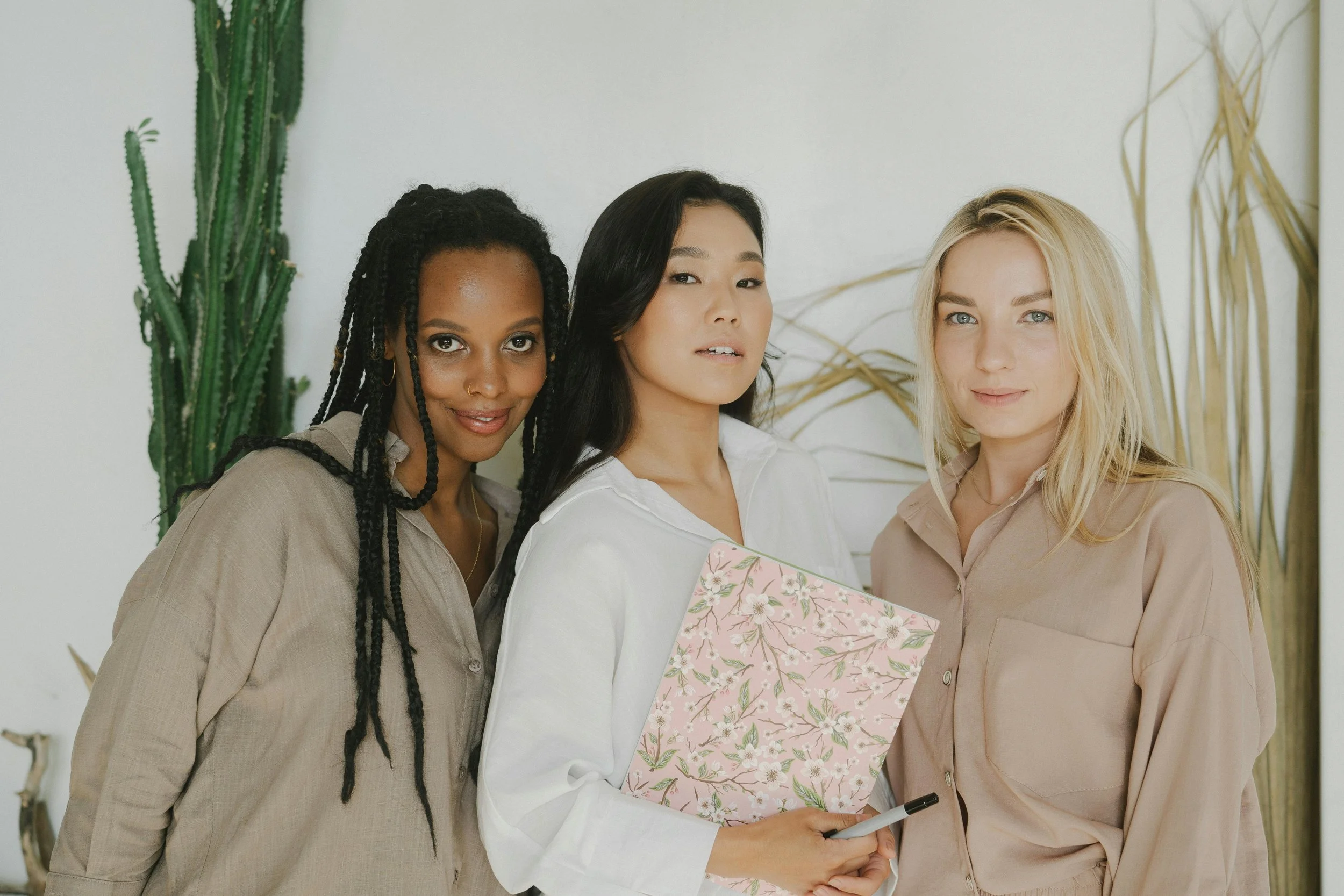 Three women standing close together indoors, one holding a pink floral-patterned folder and pen, with plants in the background.