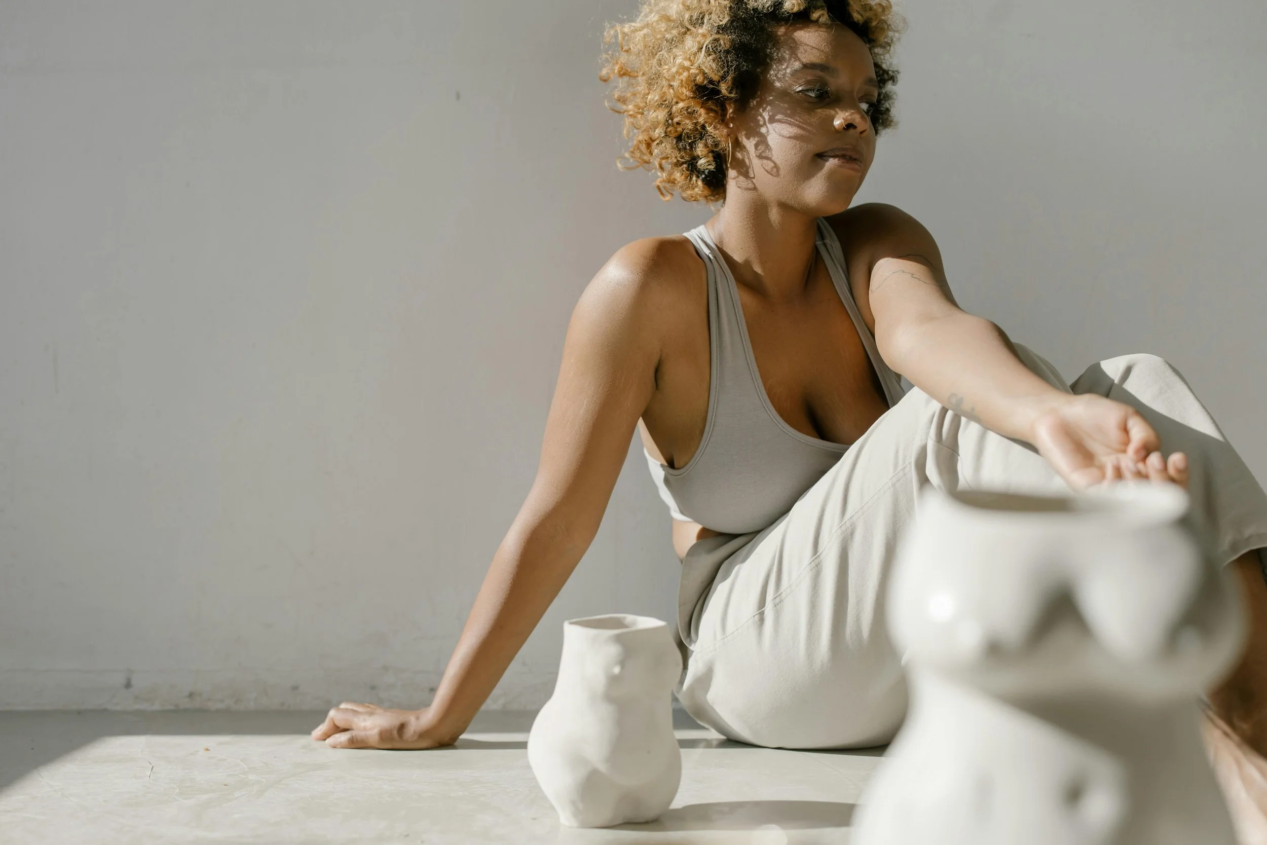 A woman with curly hair sitting on the floor in a light-colored outfit, reaching out to touch a white ceramic vase, with other vases nearby, in front of a plain background.