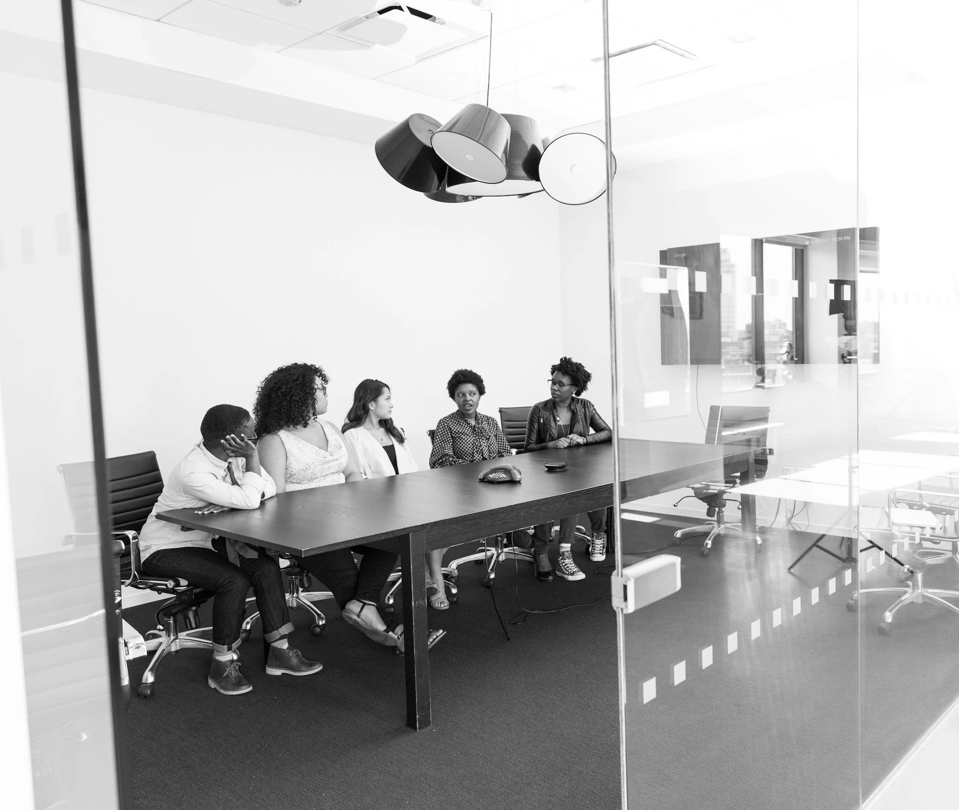 A diverse group of five women sitting at a long conference table in a modern office, engaged in a discussion.