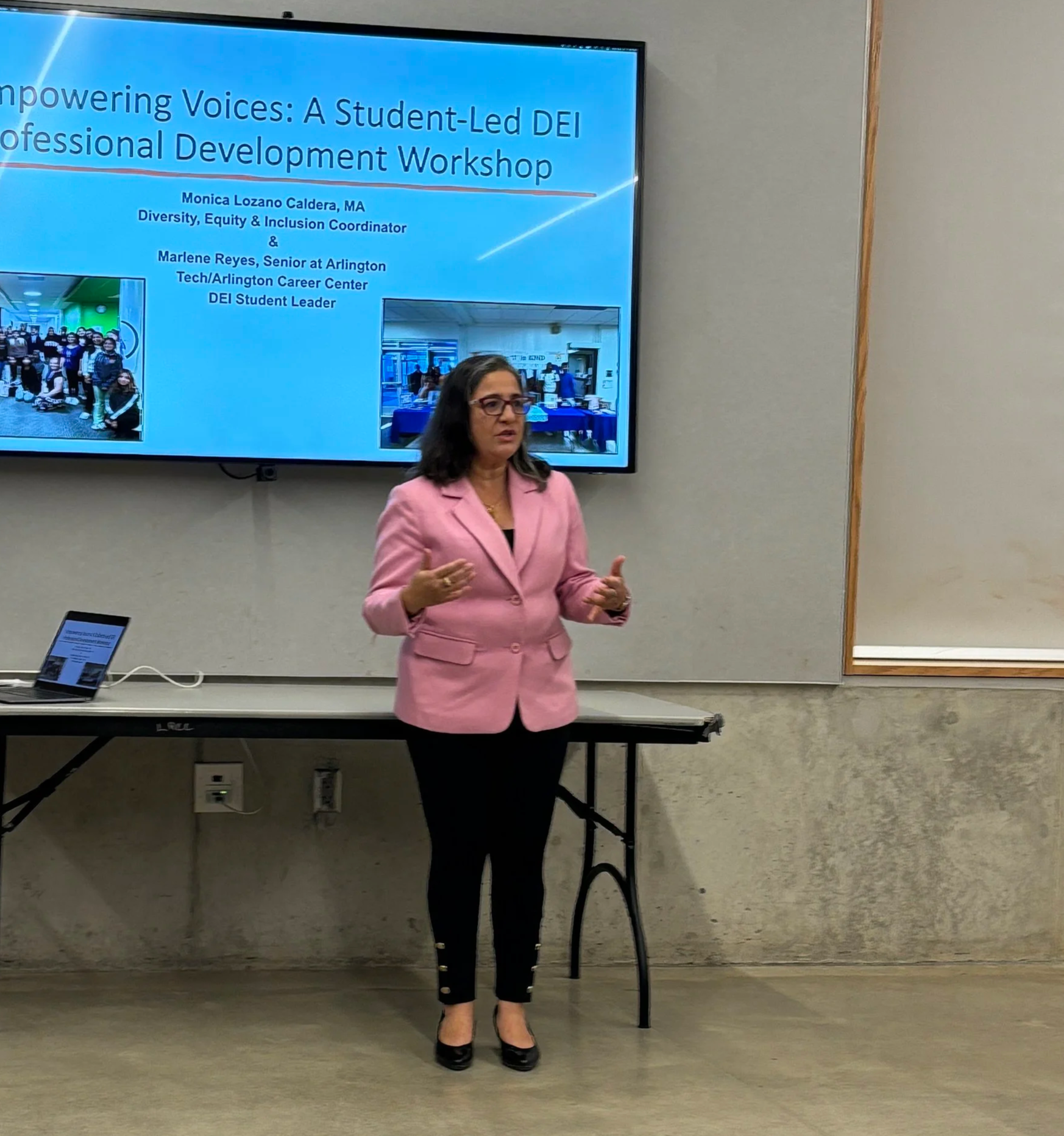 Woman wearing a pink blazer and black pants standing in front of a presentation screen with a PowerPoint slide titled "Empowering Voices: A Student-Led DEI Professional Development Workshop."