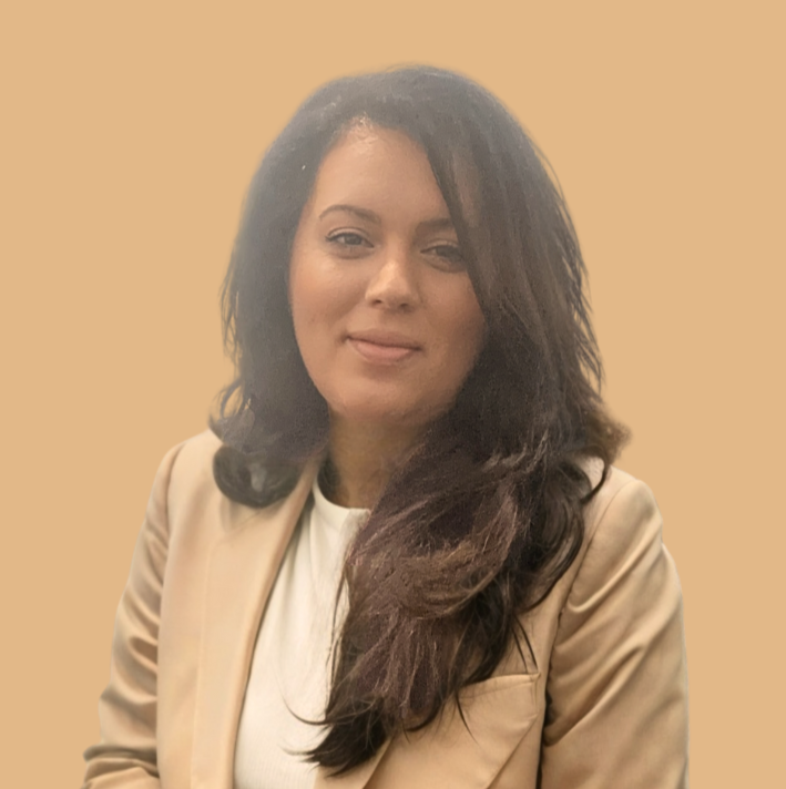 A woman with long dark hair wearing a beige blazer and white top, smiling softly against a plain beige background.