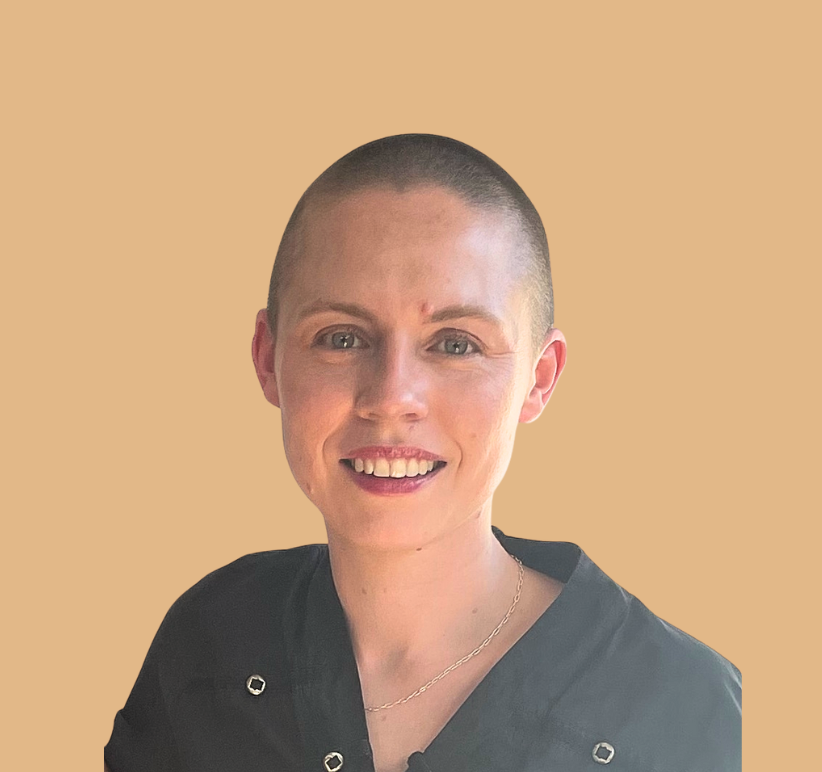 A woman with a shaved head smiling against a beige background, wearing a black top and a thin necklace.