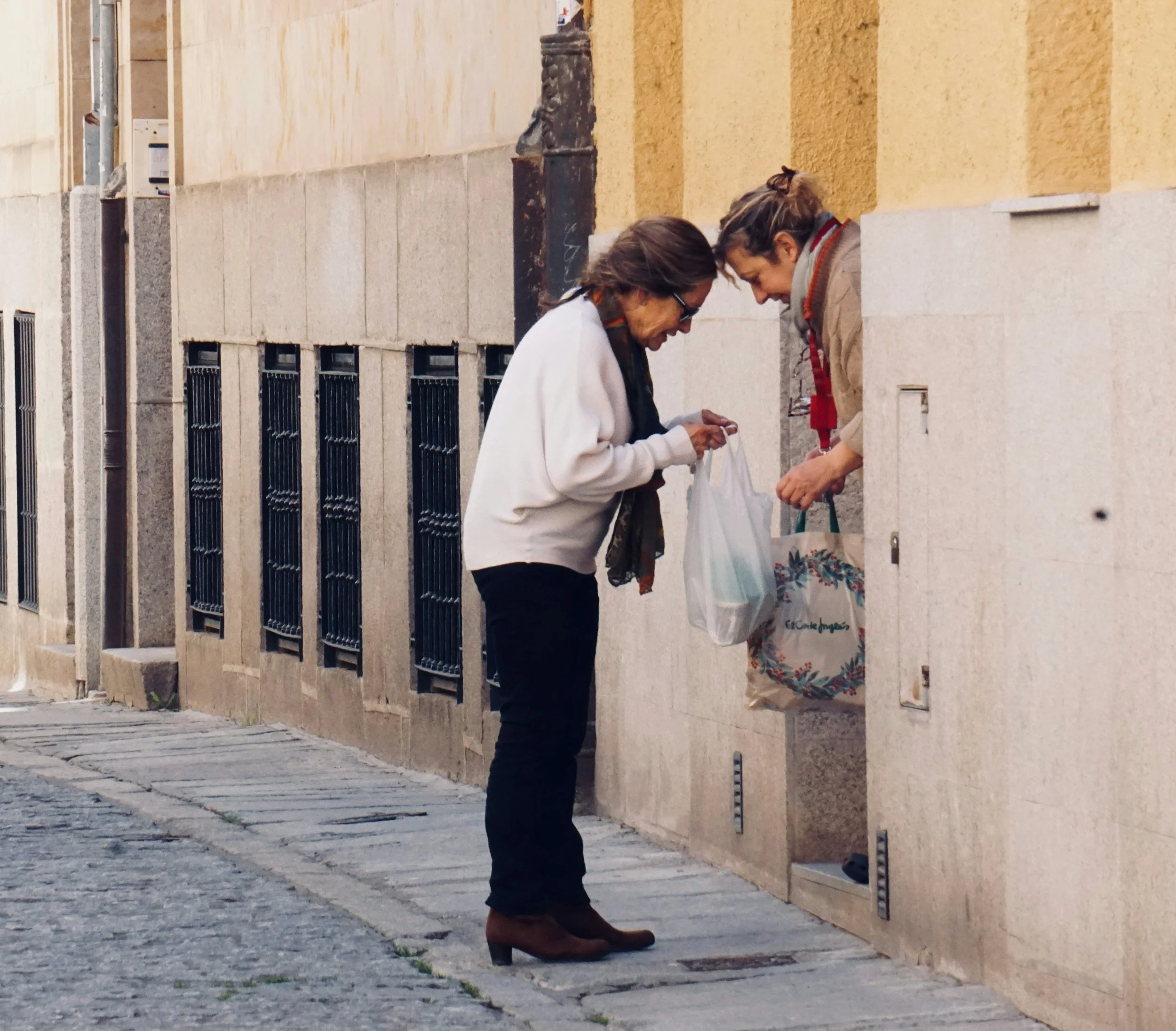Two women exchanging items at a street mailbox, standing on a cobblestone sidewalk beside a beige wall with small windows.