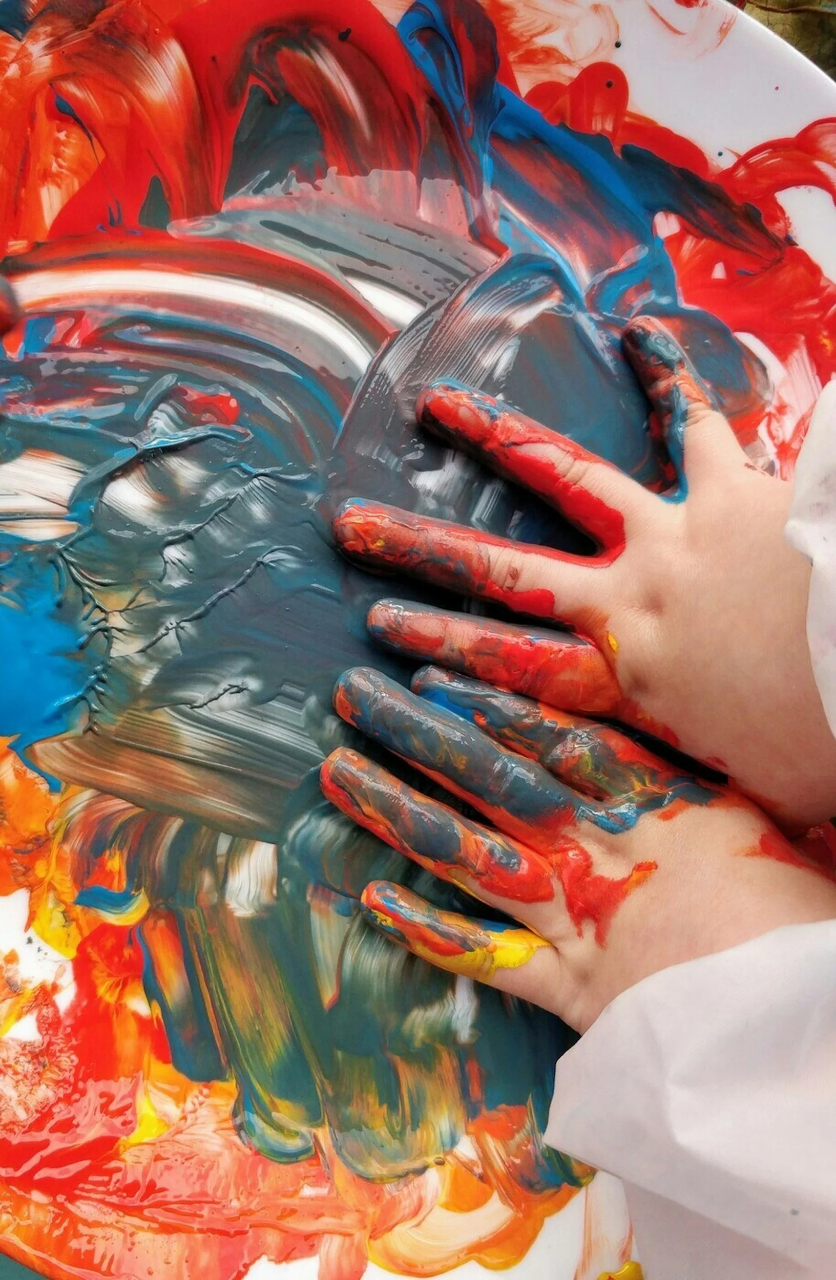 Two hands covered in multicolored paint, pressing onto a surface covered with vibrant, swirling blue, red, yellow, and white paints, creating an abstract, expressive artwork.