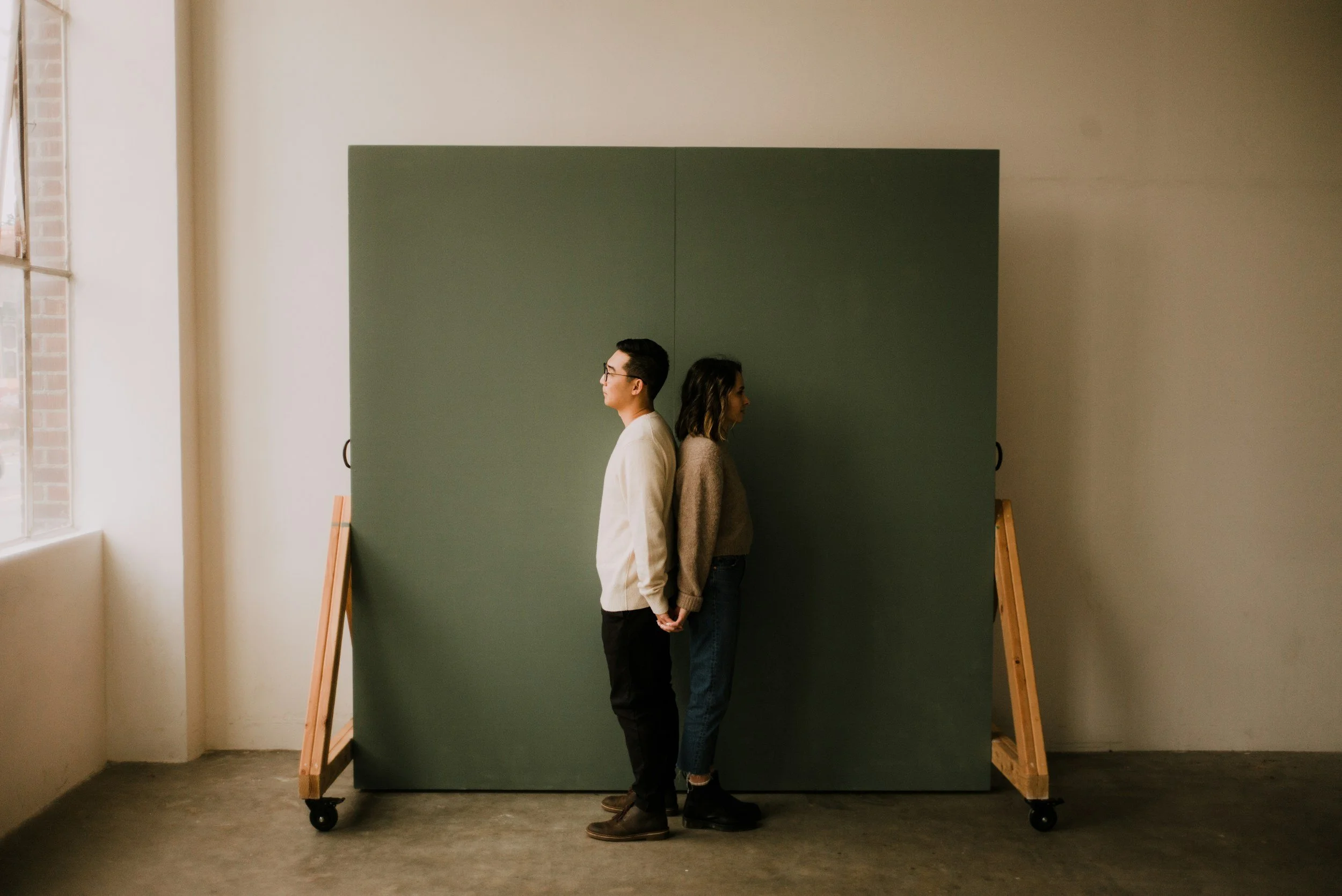 Two people standing back-to-back, holding hands, with a dark green partition behind them in a room with a window on the left side.