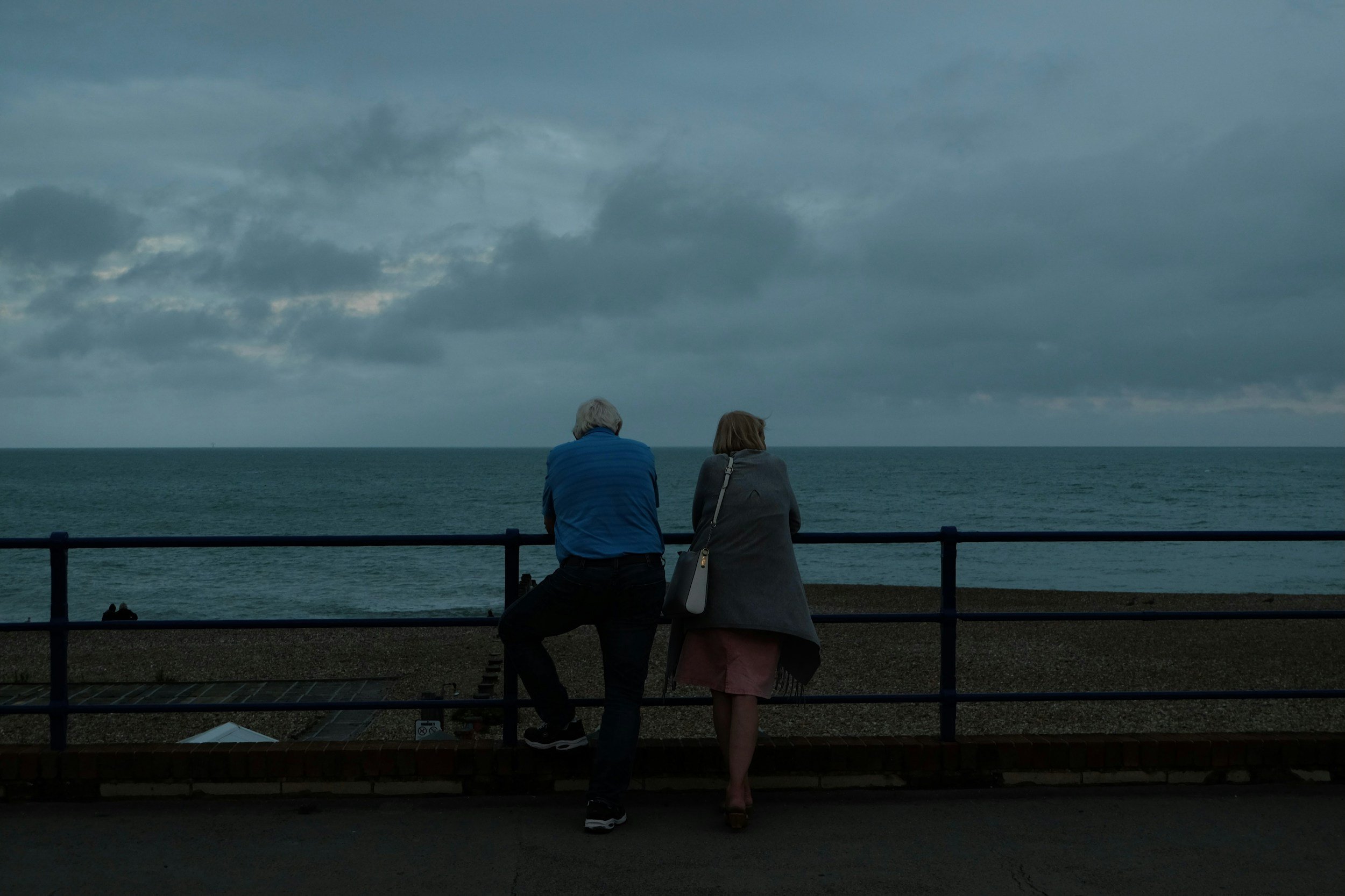 Two people, an elderly man and a woman, are sitting on a seaside promenade facing the ocean on a cloudy day.