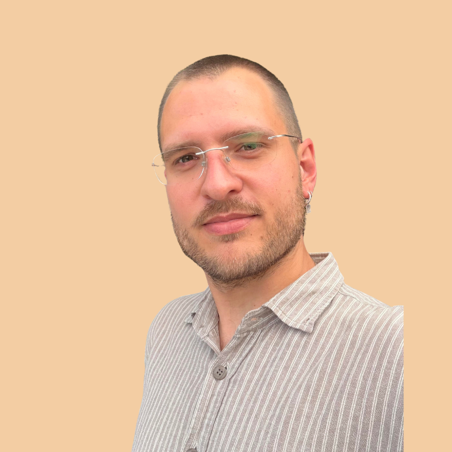 A man with short hair, beard, and glasses, wearing a striped shirt, taking a selfie against a solid beige background.