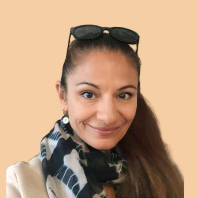 A woman with long brown hair, wearing sunglasses on her head, earrings, a patterned scarf, and a light-colored jacket, smiling at the camera against a beige background.