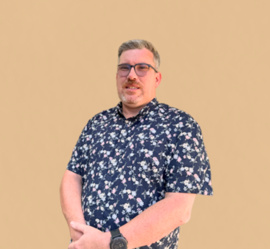 A man with glasses, short gray hair, and a beard wearing a dark floral shirt and a watch, standing against a plain beige background.