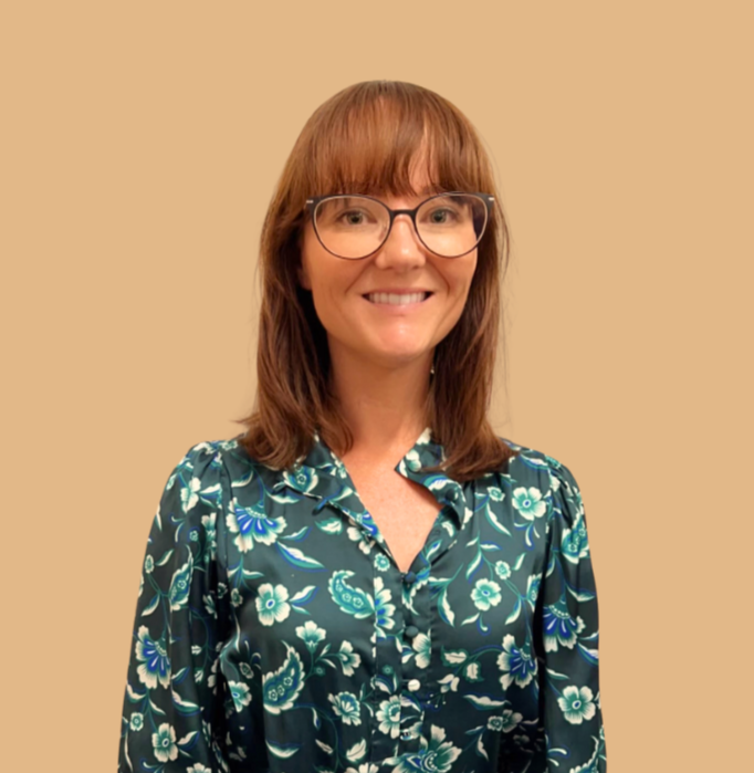 A woman with shoulder-length brown hair, glasses, and a floral-patterned shirt standing against a beige background, smiling at the camera.