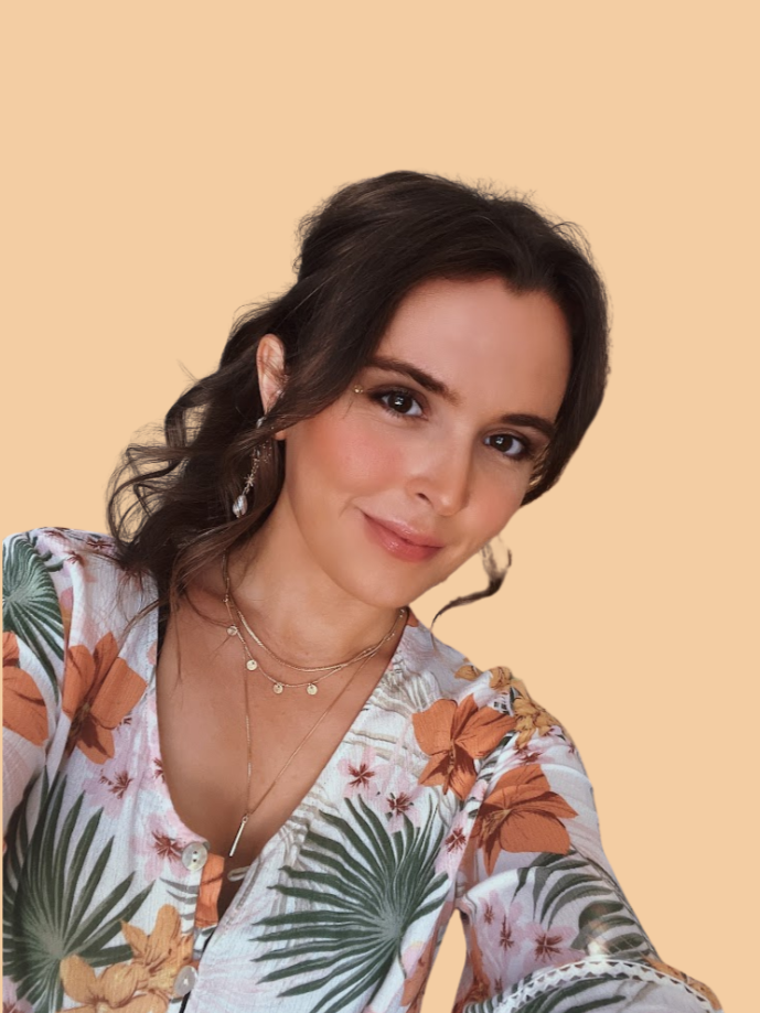 A young woman with wavy brown hair and light skin, smiling, wearing a floral-patterned shirt with orange and green leaves, layered necklaces, and drop earrings, against a beige background.