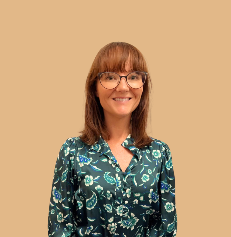 A woman with shoulder-length auburn hair, glasses, and a floral satin shirt, smiling against a beige background.