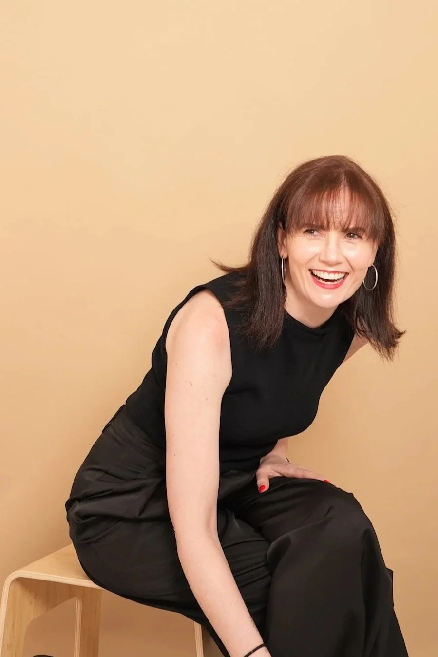 A woman with shoulder-length brown hair, wearing a black sleeveless top and black pants, sitting on a light-colored wooden stool against a beige background, smiling broadly.