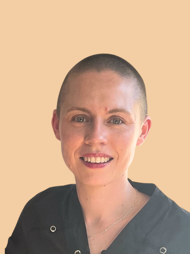 A woman with a shaved head smiling at the camera, wearing a black top with silver buttons and a necklace, against a beige background.