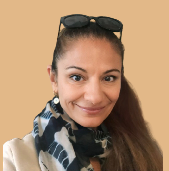 A woman with dark hair tied back, wearing sunglasses on her head, a beige top, and a black and white patterned scarf, smiling against a plain beige background.