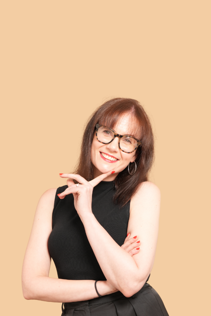 A woman with shoulder-length brown hair, wearing glasses and hoop earrings, smiling, with one hand resting on her chin and the other arm crossed, against a beige background.