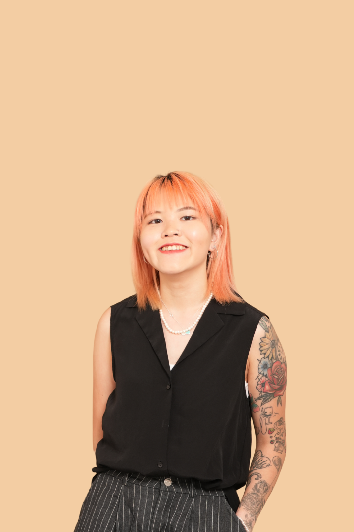 Young woman with orange hair and tattoos, wearing a black sleeveless blouse, striped pants, and a pearl necklace, smiling against a beige background.