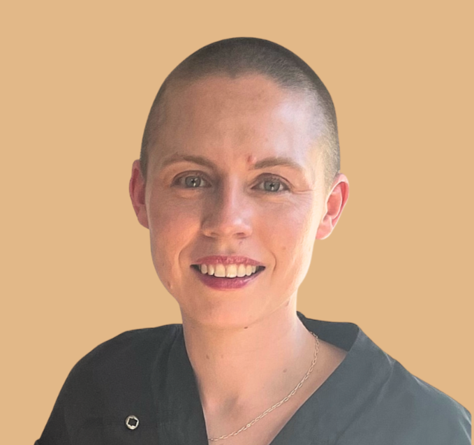 A woman with a shaved head, smiling in front of a beige background, wearing a dark top and a delicate chain necklace.