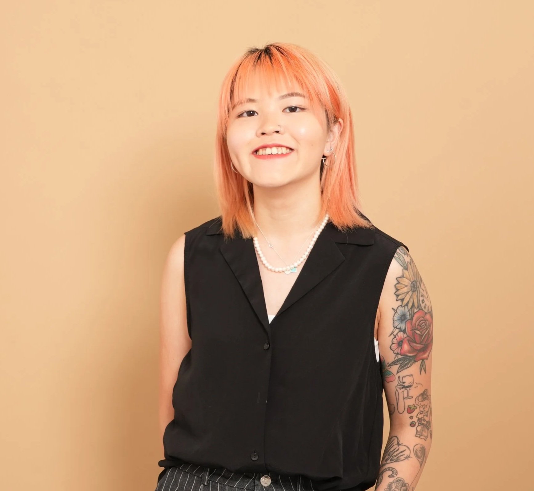 A smiling woman with peach-colored hair wearing a sleeveless black shirt, pearl necklace, and earrings, standing against a beige background with tattoos on her left arm.