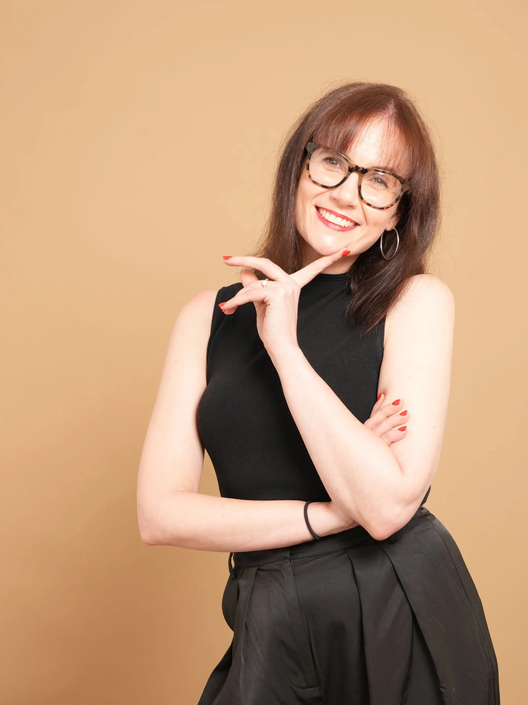 A woman with shoulder-length brown hair, glasses, and hoop earrings, smiling, wearing a sleeveless black top and black pants, standing against a plain beige background.