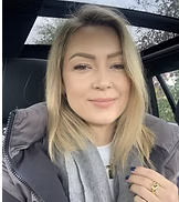 A young woman with blonde hair smiling inside a car, wearing a gray jacket.