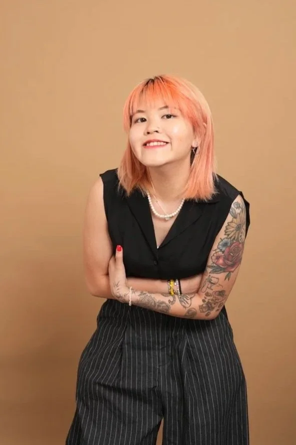 A woman with pink hair smiling, wearing a black sleeveless top, pinstripe pants, and jewelry, standing against a beige background.