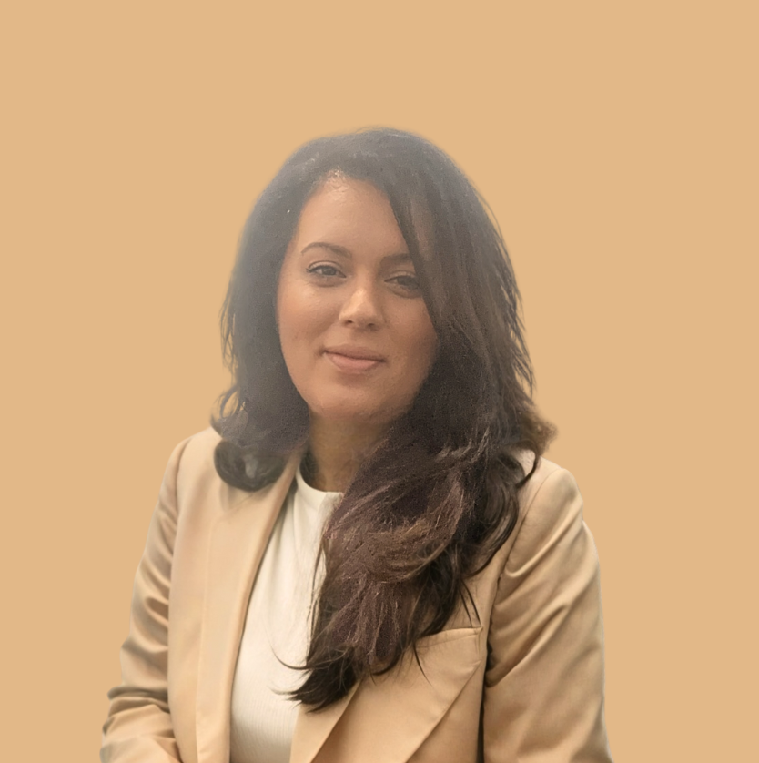 A woman with long dark hair wearing a beige blazer and white top, standing against a plain beige background.