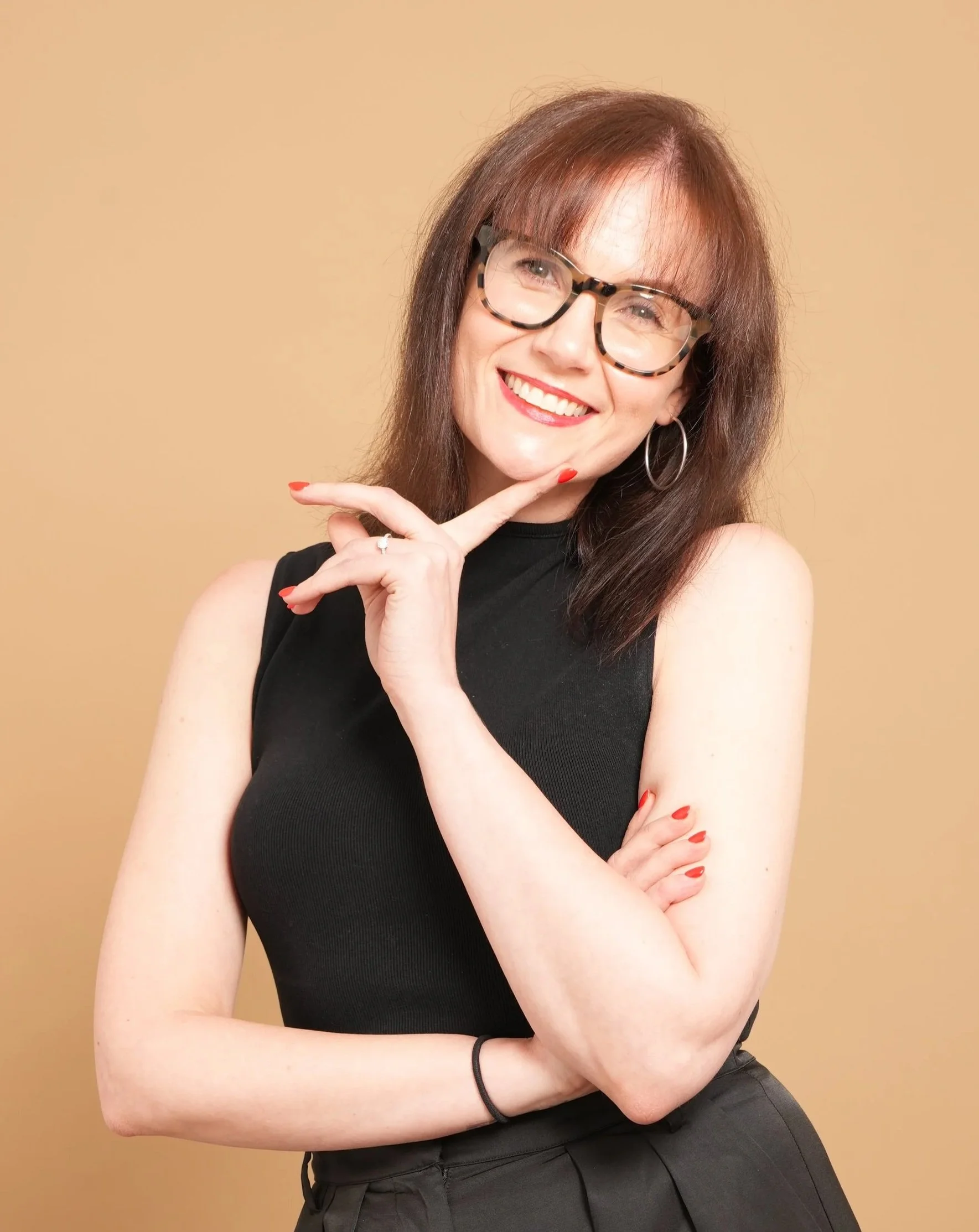 Smiling woman with brown hair, glasses, in a black sleeveless top, posing with her hand on her chin against a beige background.