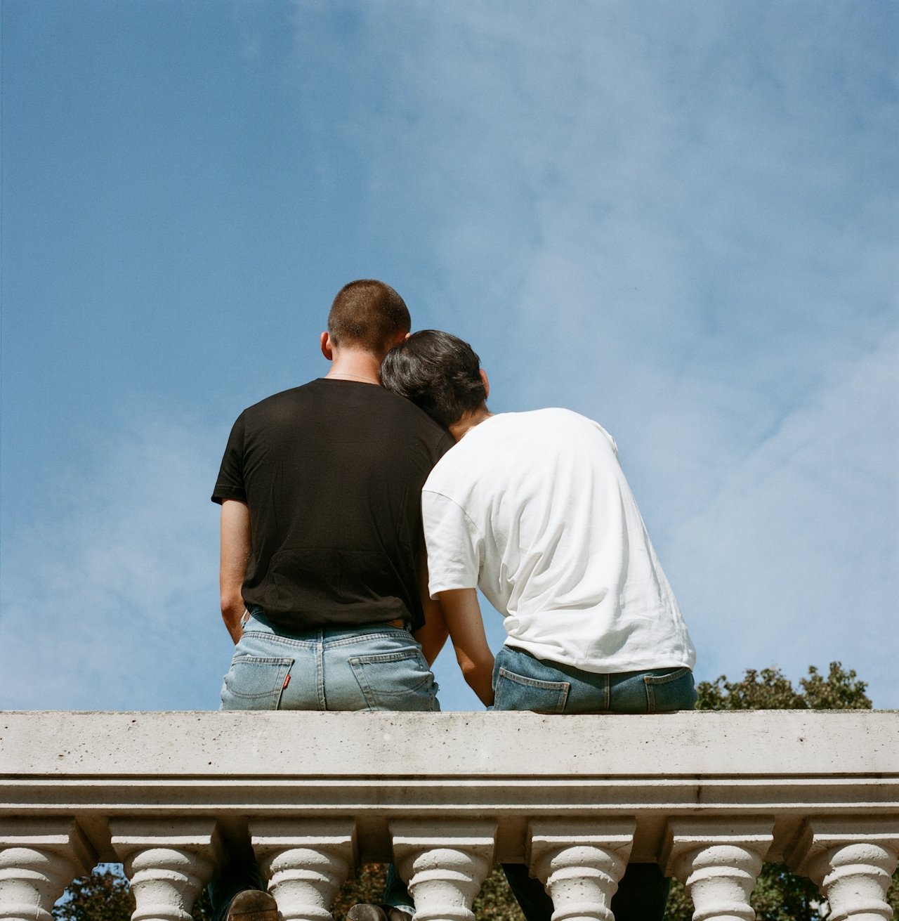 Two people sitting side by side on a stone railing, with their heads touching, against a blue sky background. One is wearing a black shirt and the other a white shirt.