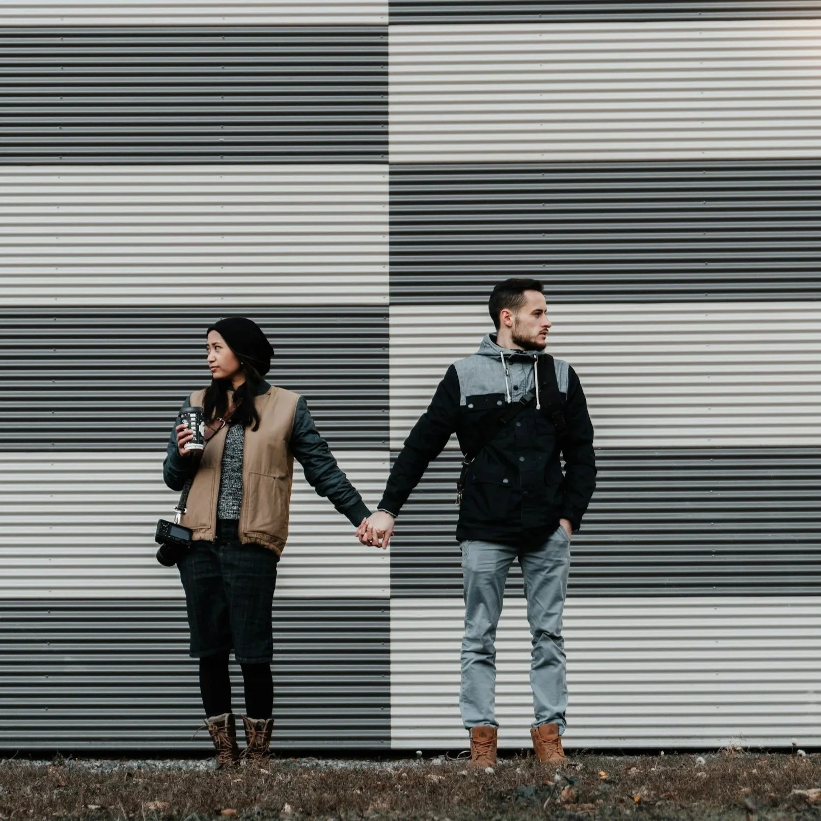 A couple holding hands stands in front of a large, geometric, black-and-white striped wall. The woman holds a coffee cup and wears a black beanie, tan vest, black pants, and brown boots. The man looks to the right, wears a gray and black jacket, light gray pants, and brown boots.