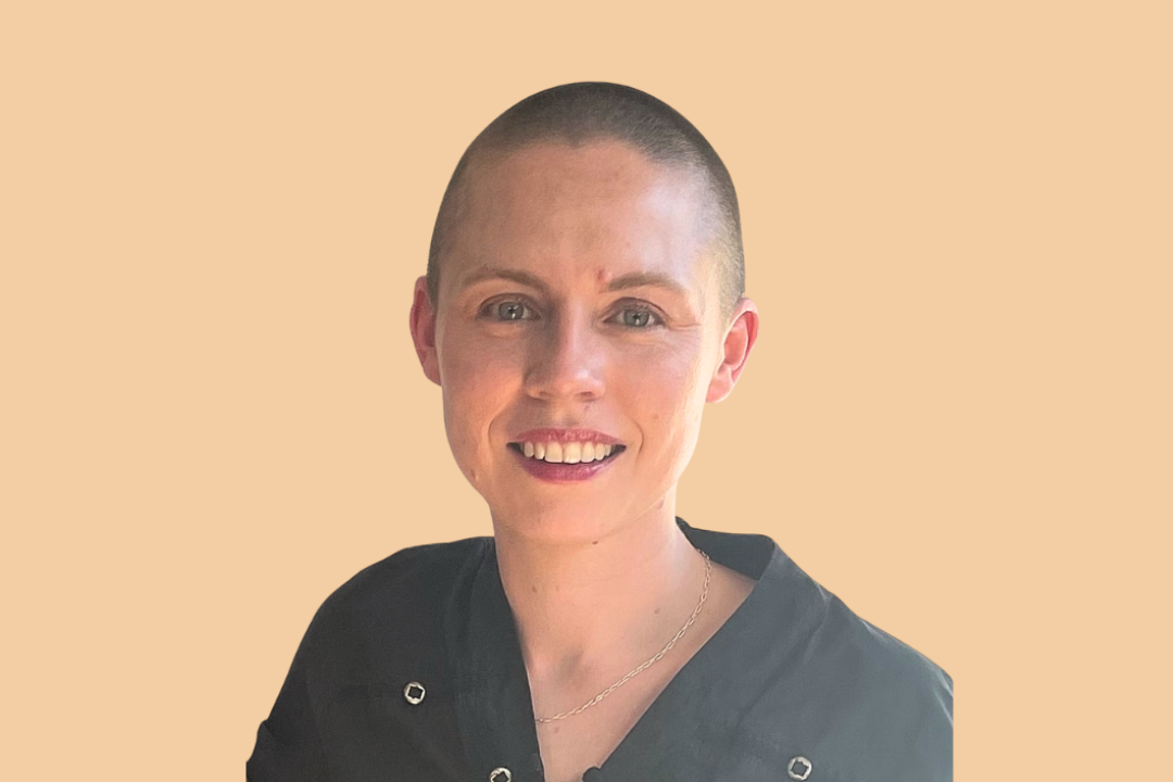 Close-up of a woman with a buzz cut, smiling, wearing a black top with metallic buttons and a thin gold necklace, against a beige background.