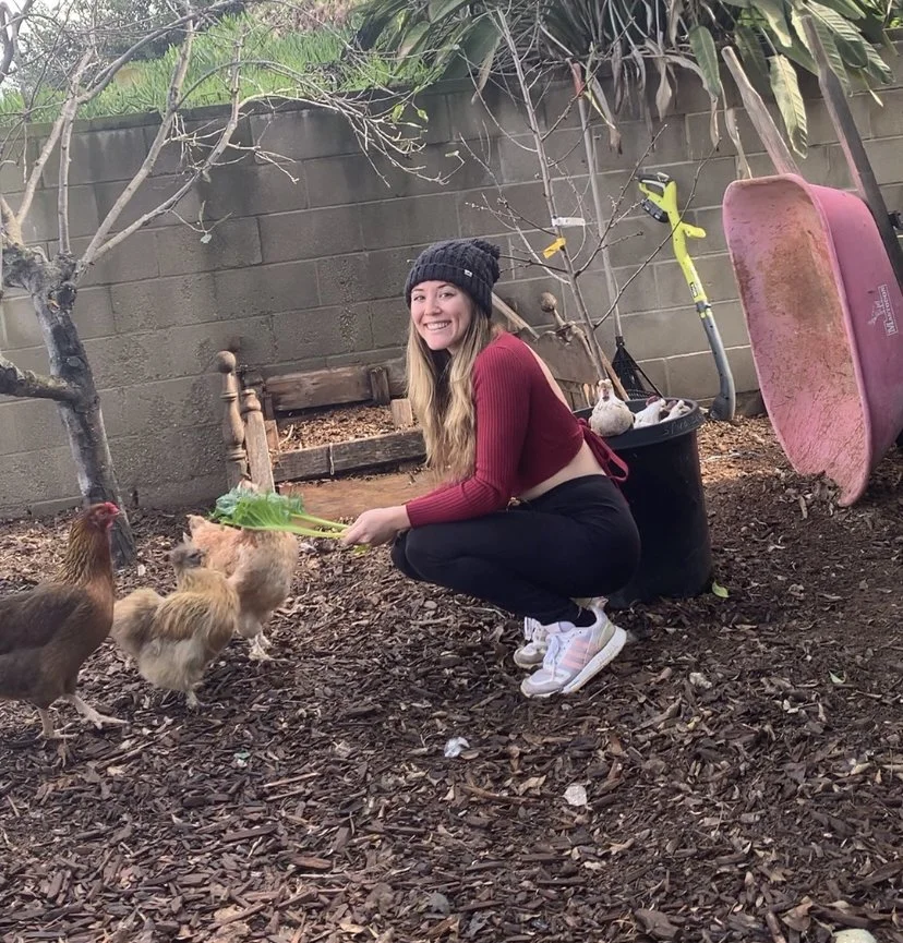 A young woman with long hair, wearing a black beanie, and a red long sleeve shirt, squatting in a backyard, feeding chickens with leafy greens. She is smiling, and there is a tree, a wooden bench, a large black planter with gardening tools, and a pin