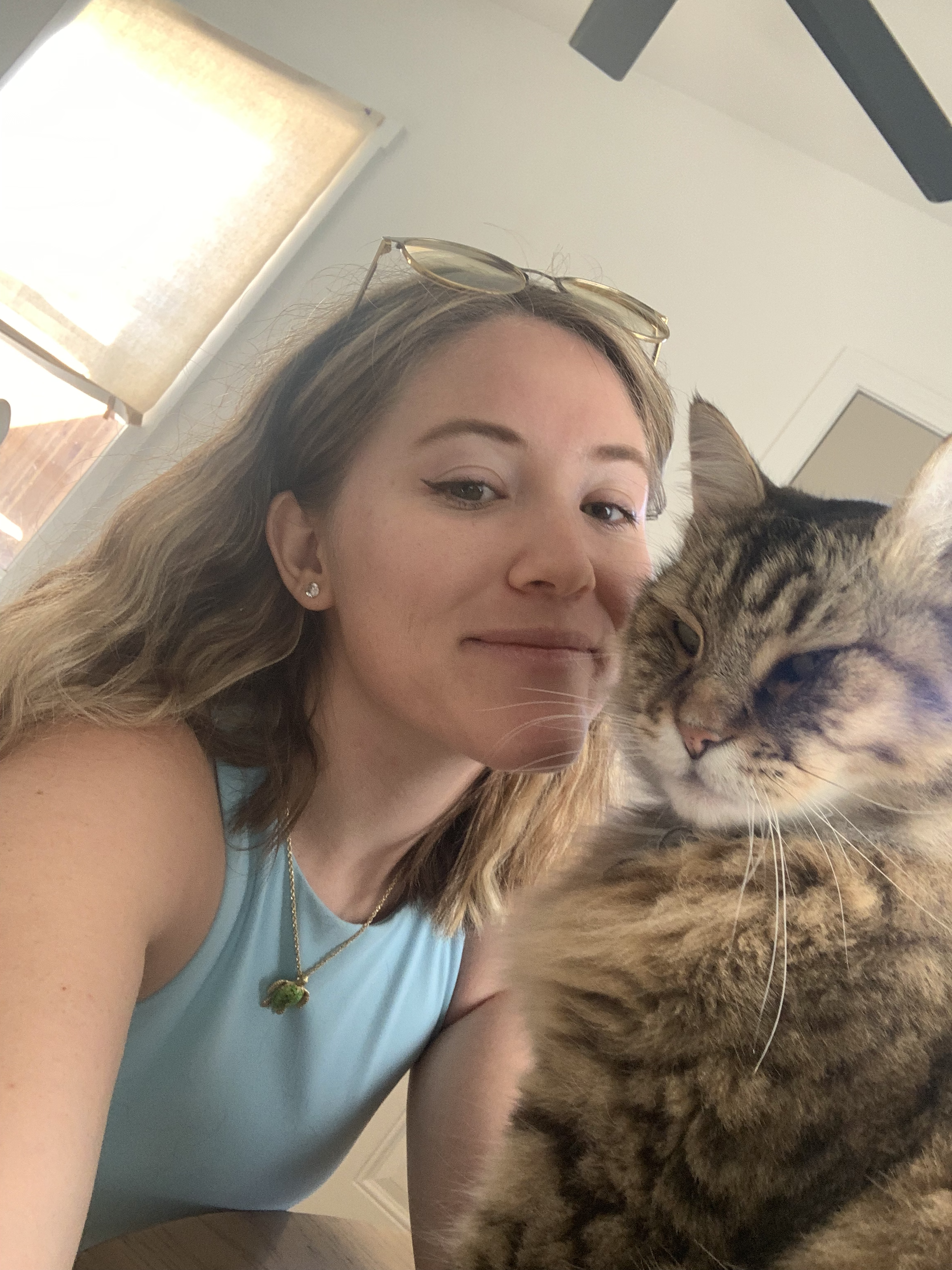 A young woman with blonde hair, wearing a light blue tank top and sunglasses on her head, taking a selfie with a large tabby cat showing a relaxed expression.