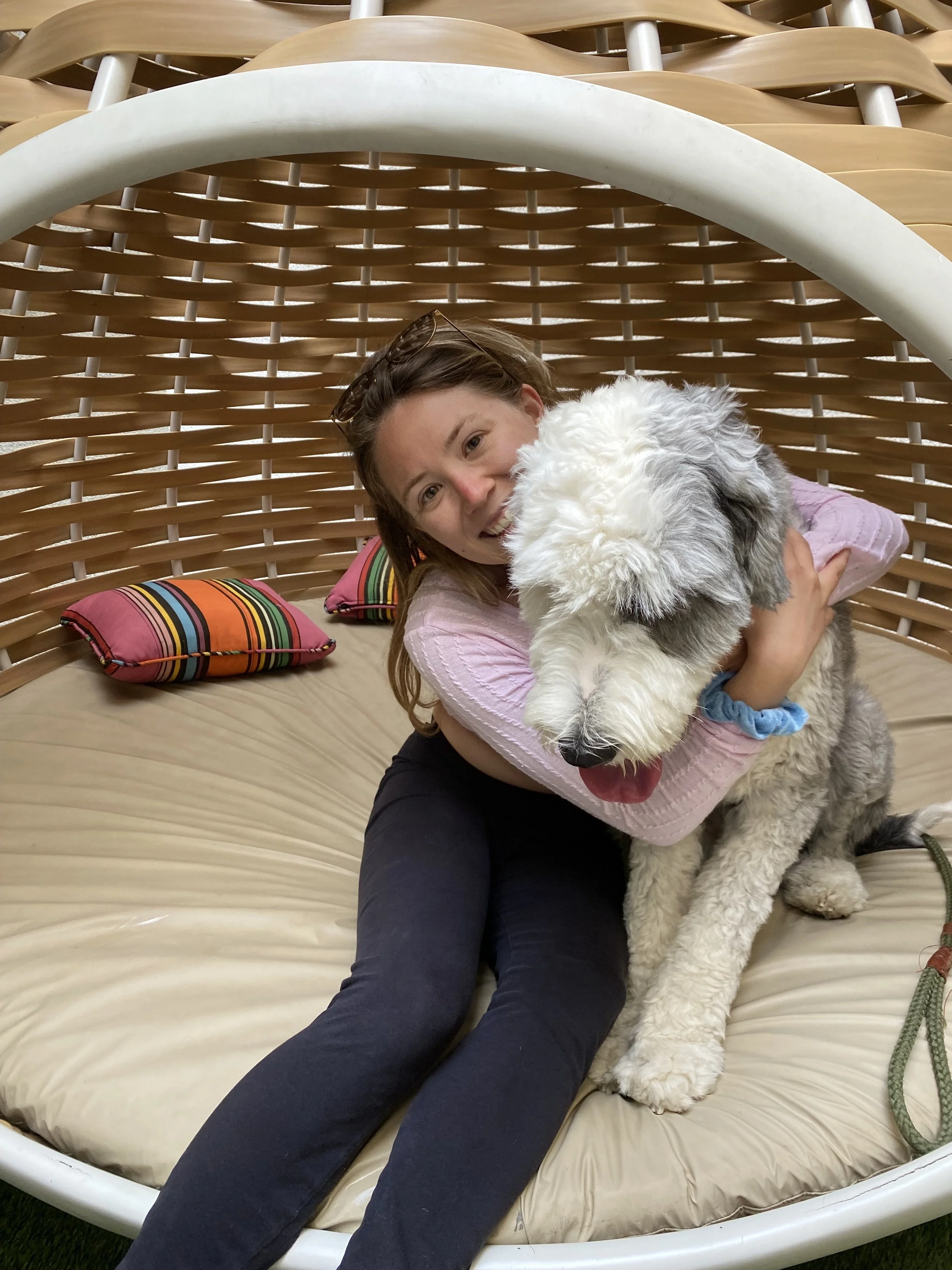 A woman and a fluffy dog sitting together on a round wicker-style outdoor daybed with striped pillows.