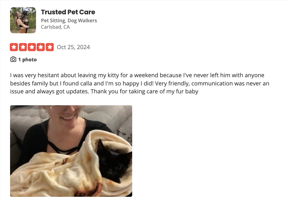 A woman smiles as she holds a black kitten wrapped in a cream-colored blanket, sitting on a gray couch.