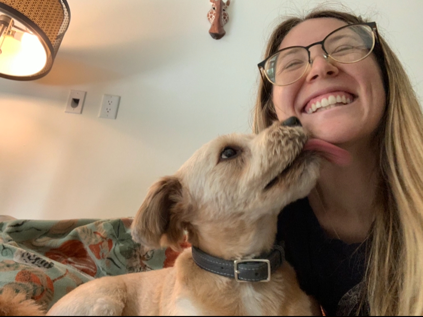 A woman with glasses smiling as a puppy licks her face indoors.