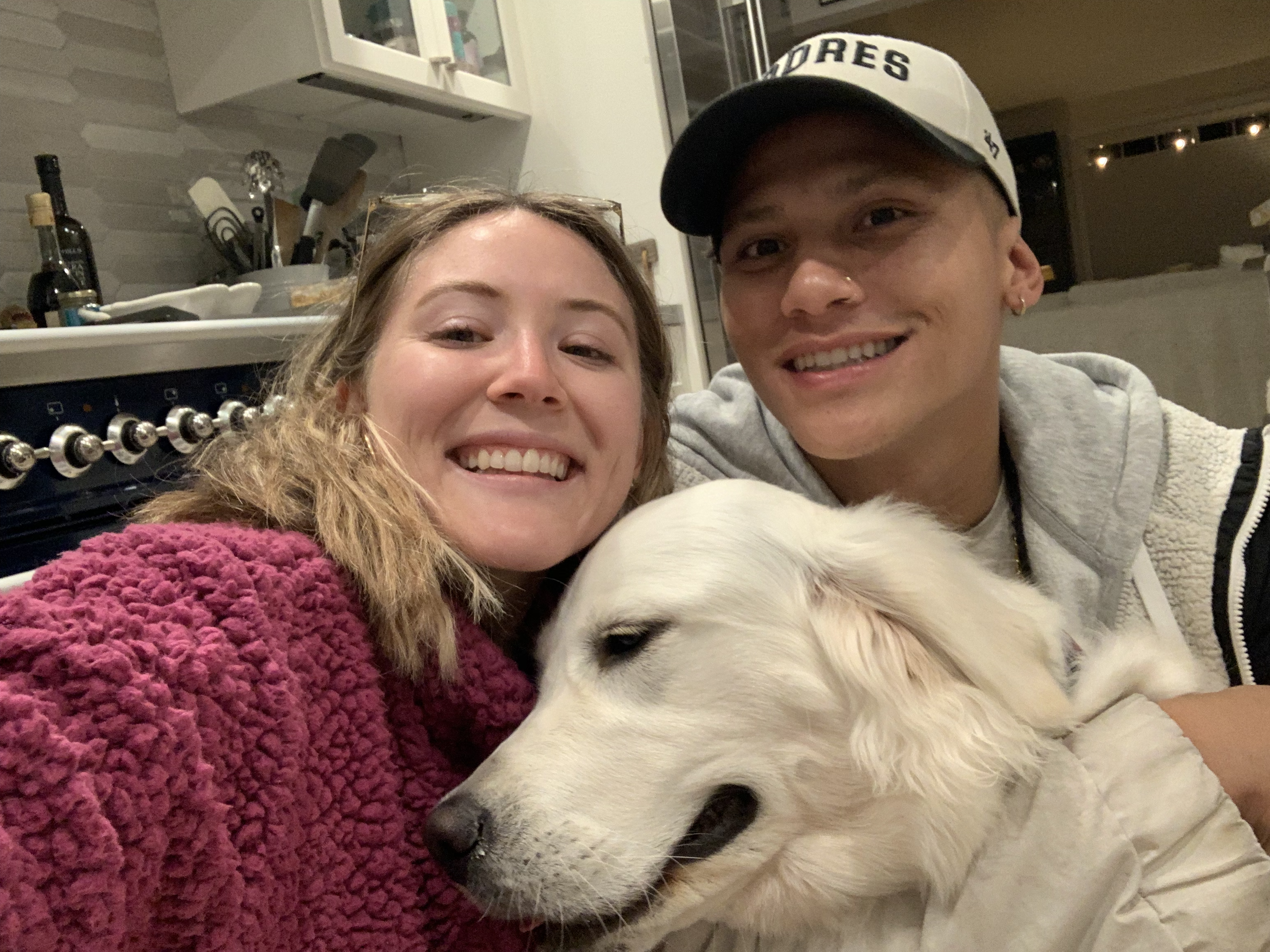 Two smiling people, a woman with curly hair wearing a pink sweater and a man with a cap, are cuddling a sleeping white dog with floppy ears.