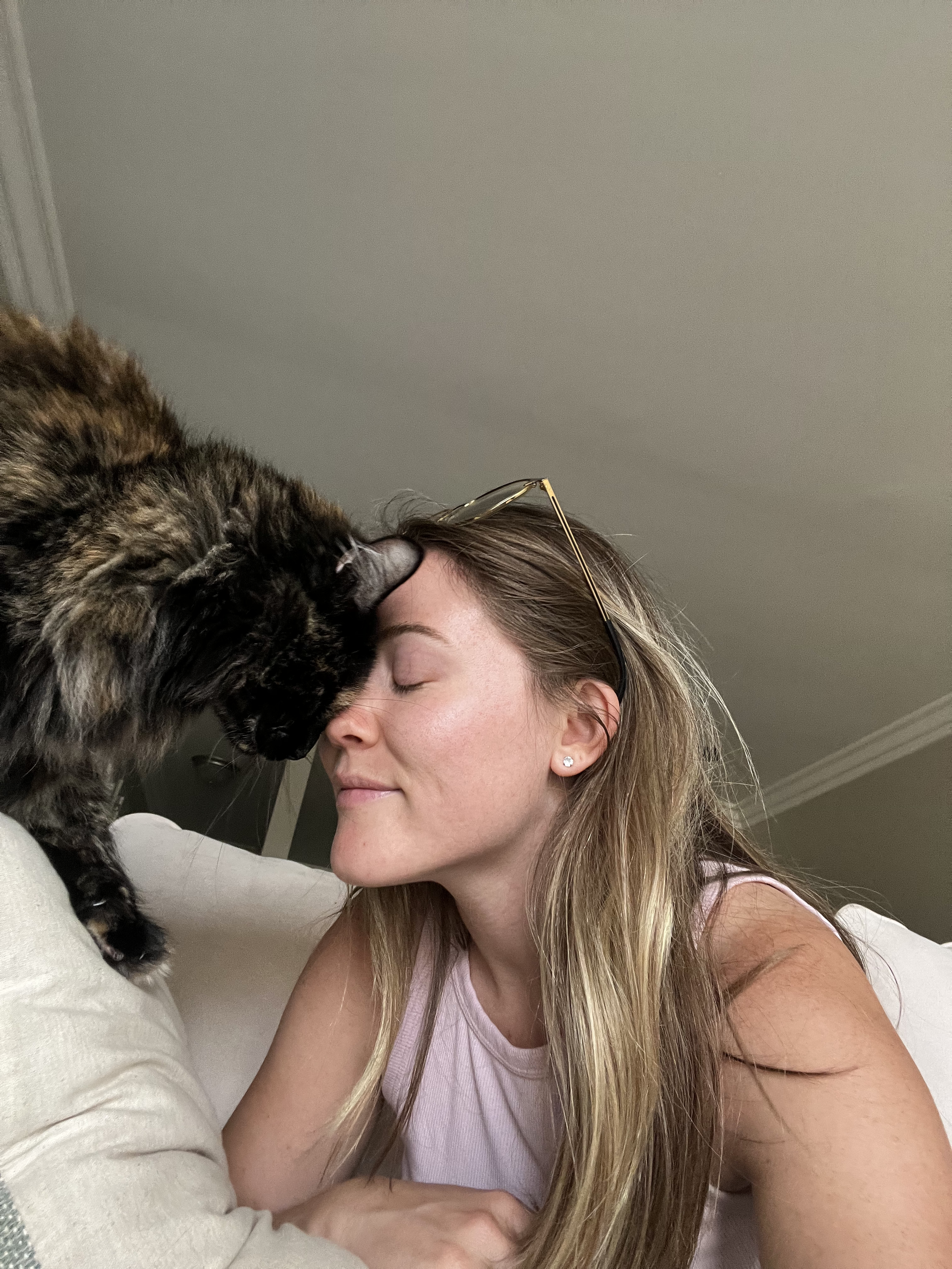 A woman with long blonde hair and earrings smiles with her eyes closed while a fluffy, dark-colored cat nuzzles her forehead.