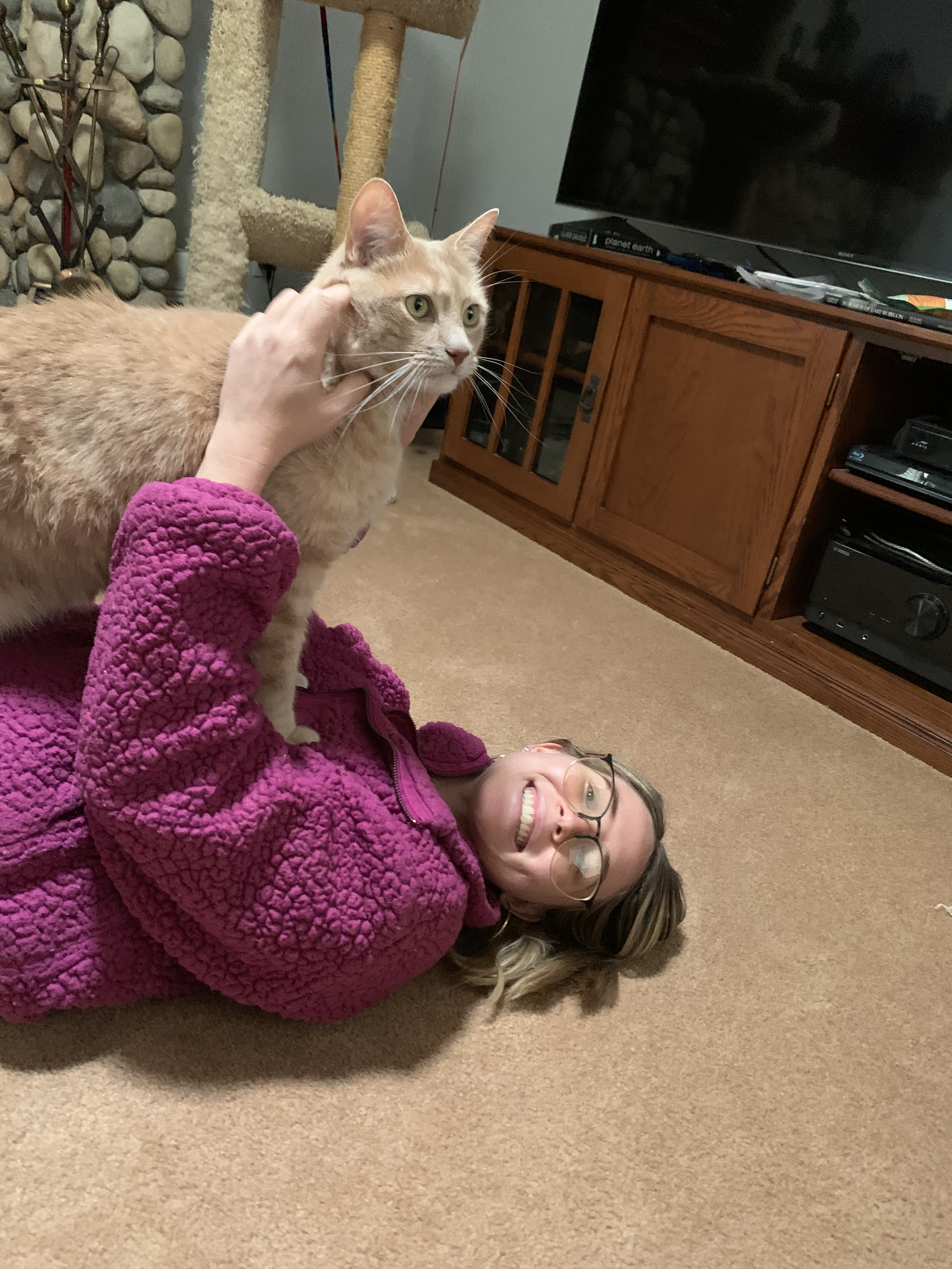 A woman lying on the carpeted floor, smiling and wearing glasses and a pink fleece jacket, is petting a ginger cat.