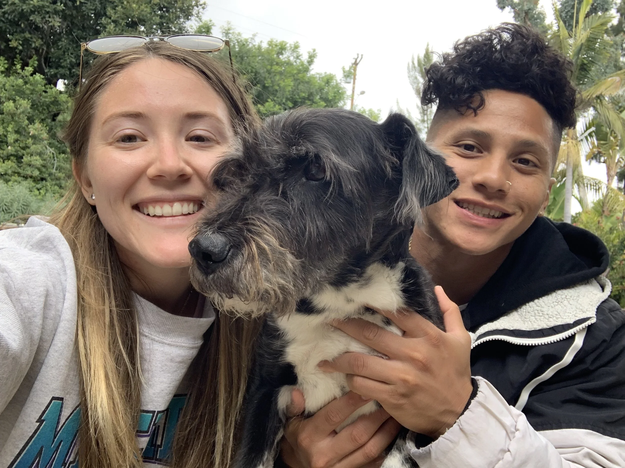 Two smiling women hold a black and white dog outdoors with greenery and trees in the background.