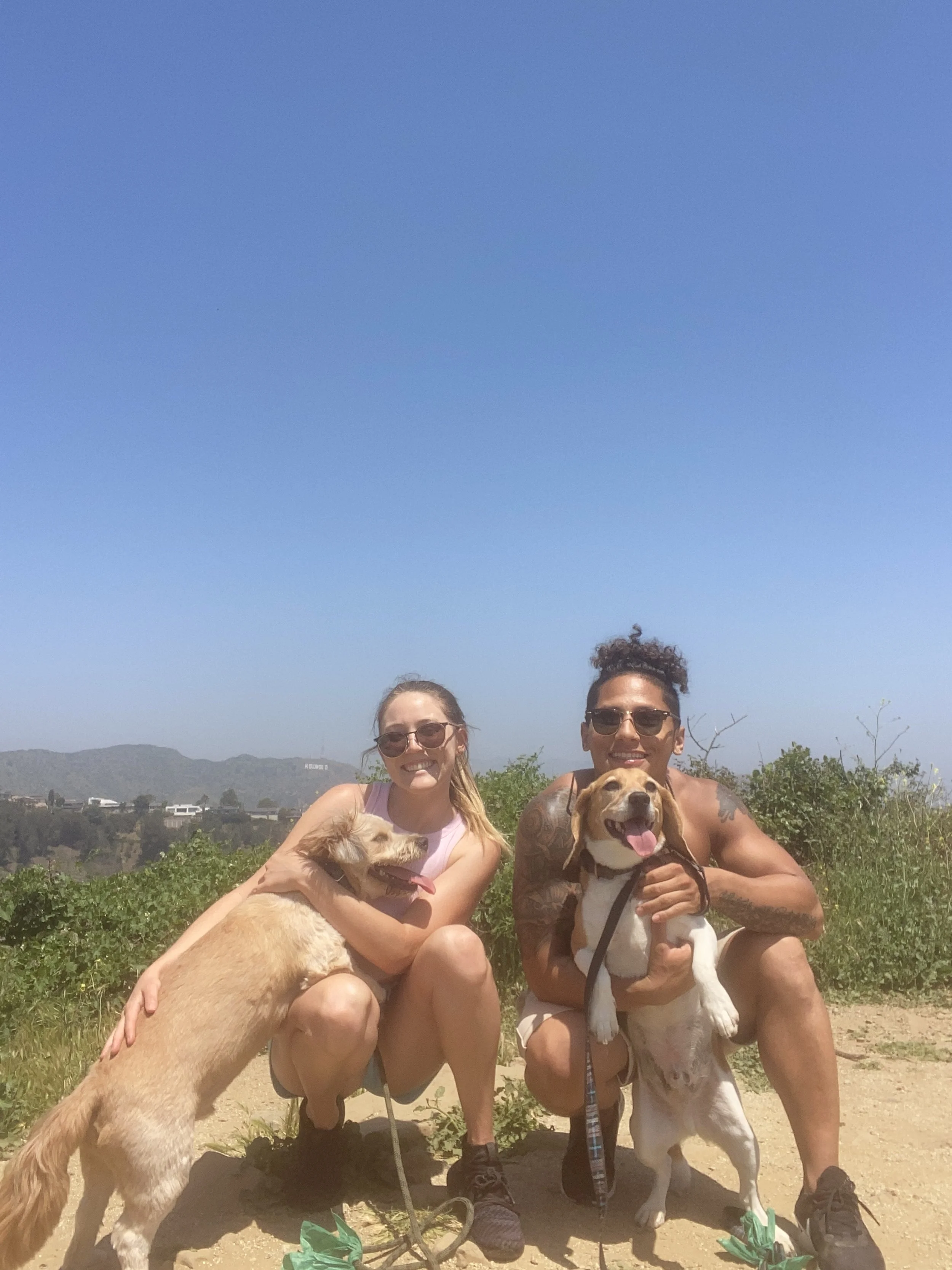 Two people, a woman and a man, crouching outdoors on a trail with greenery and hills in the background, each holding a happy dog, all wearing sunglasses and smiling on a sunny day.