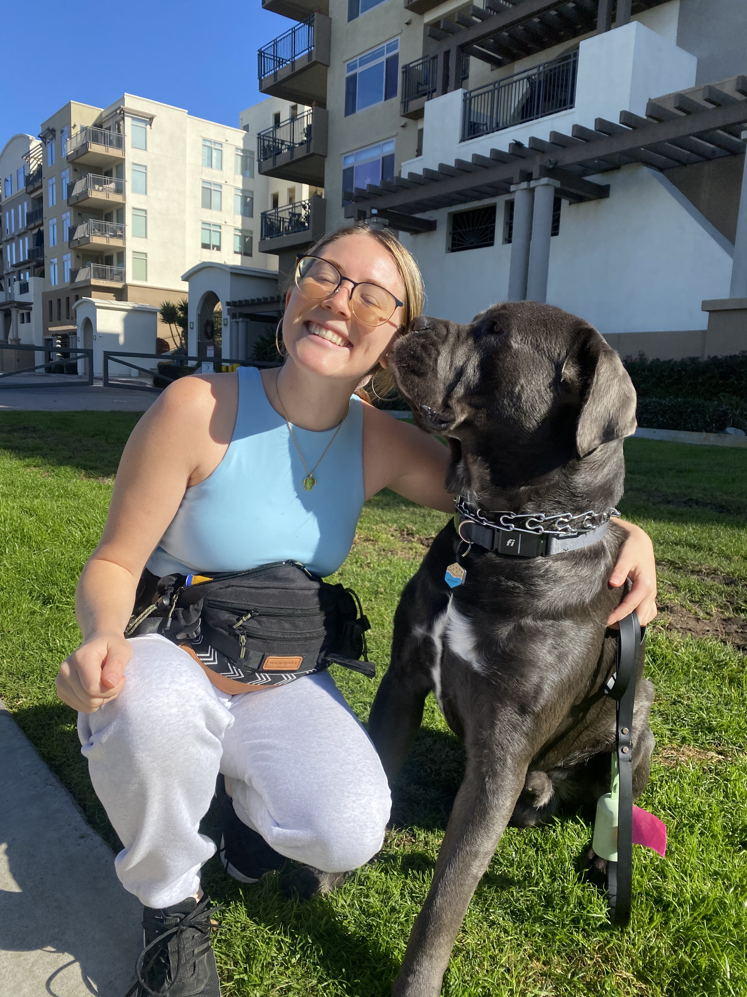 A young woman with glasses and a big smile kneeling on the grass next to a large black Great Dane dog, both looking happy. The woman is wearing a blue sleeveless top, white sweatpants, and black sneakers, with a waist bag around her waist. The backgr