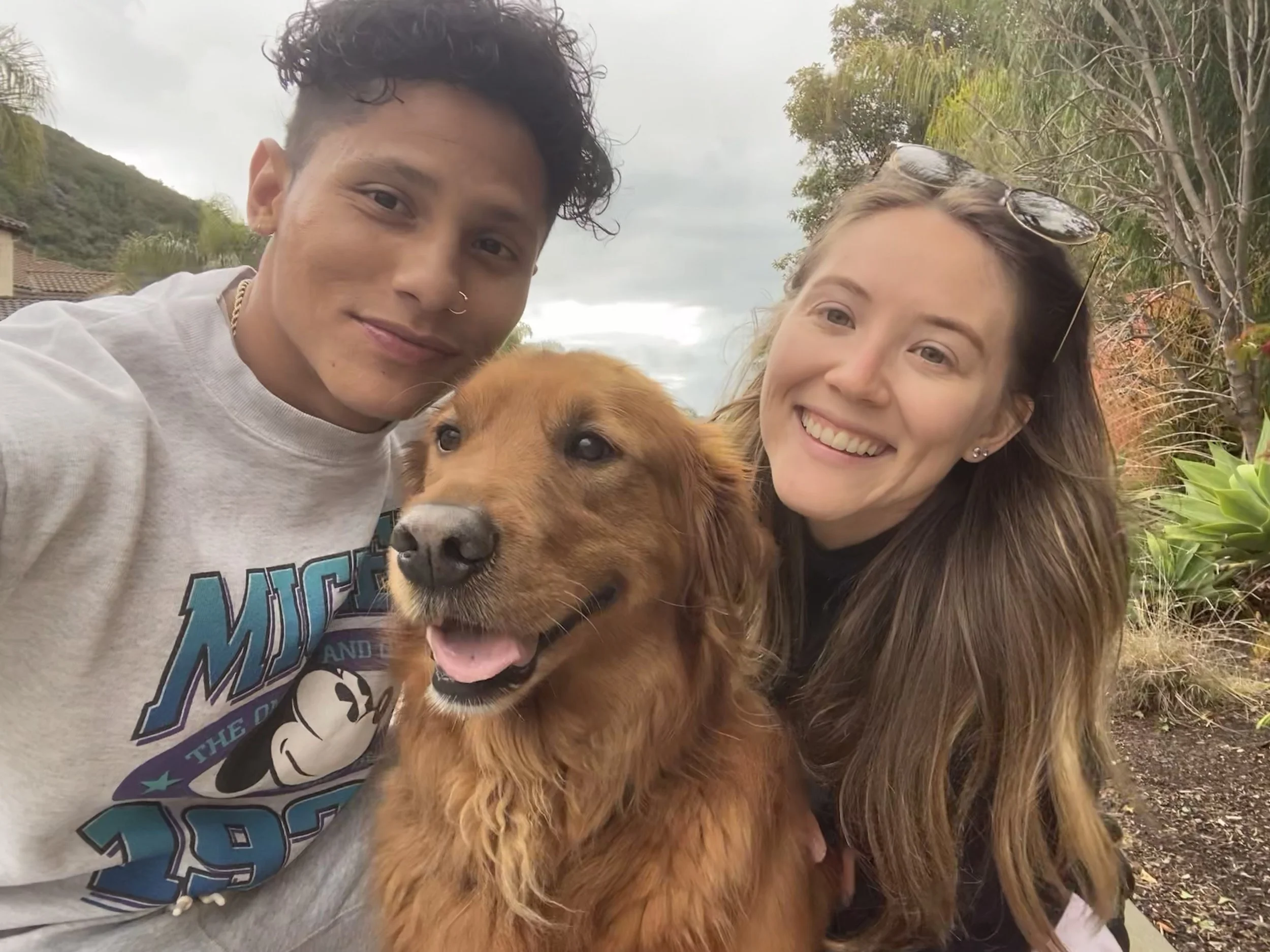 Two women and a man with a golden retriever dog outdoors in a garden or park area.