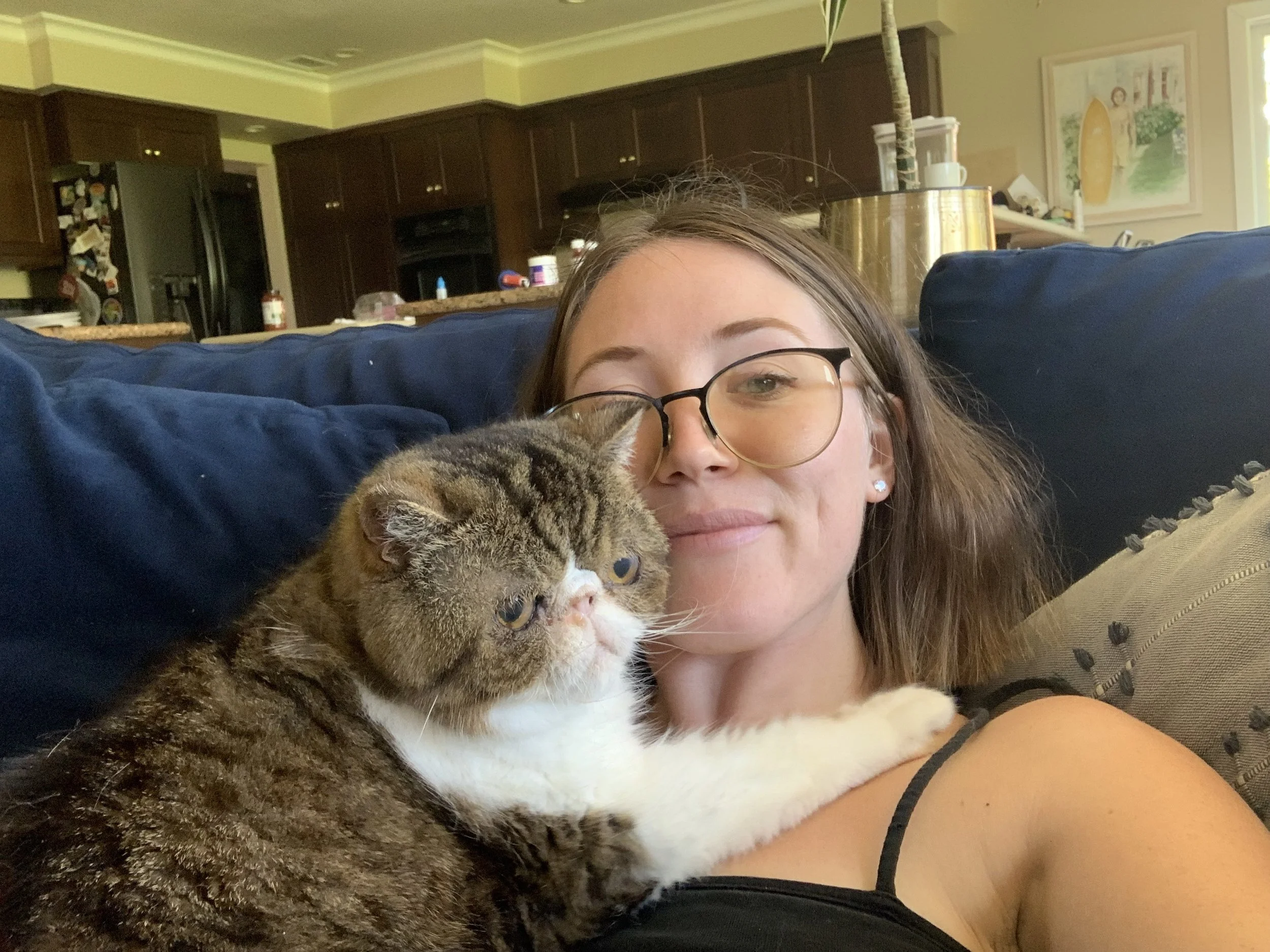 Young woman with glasses lying on a couch, holding a grumpy-looking tabby cat, in a cozy home with a kitchen in the background.