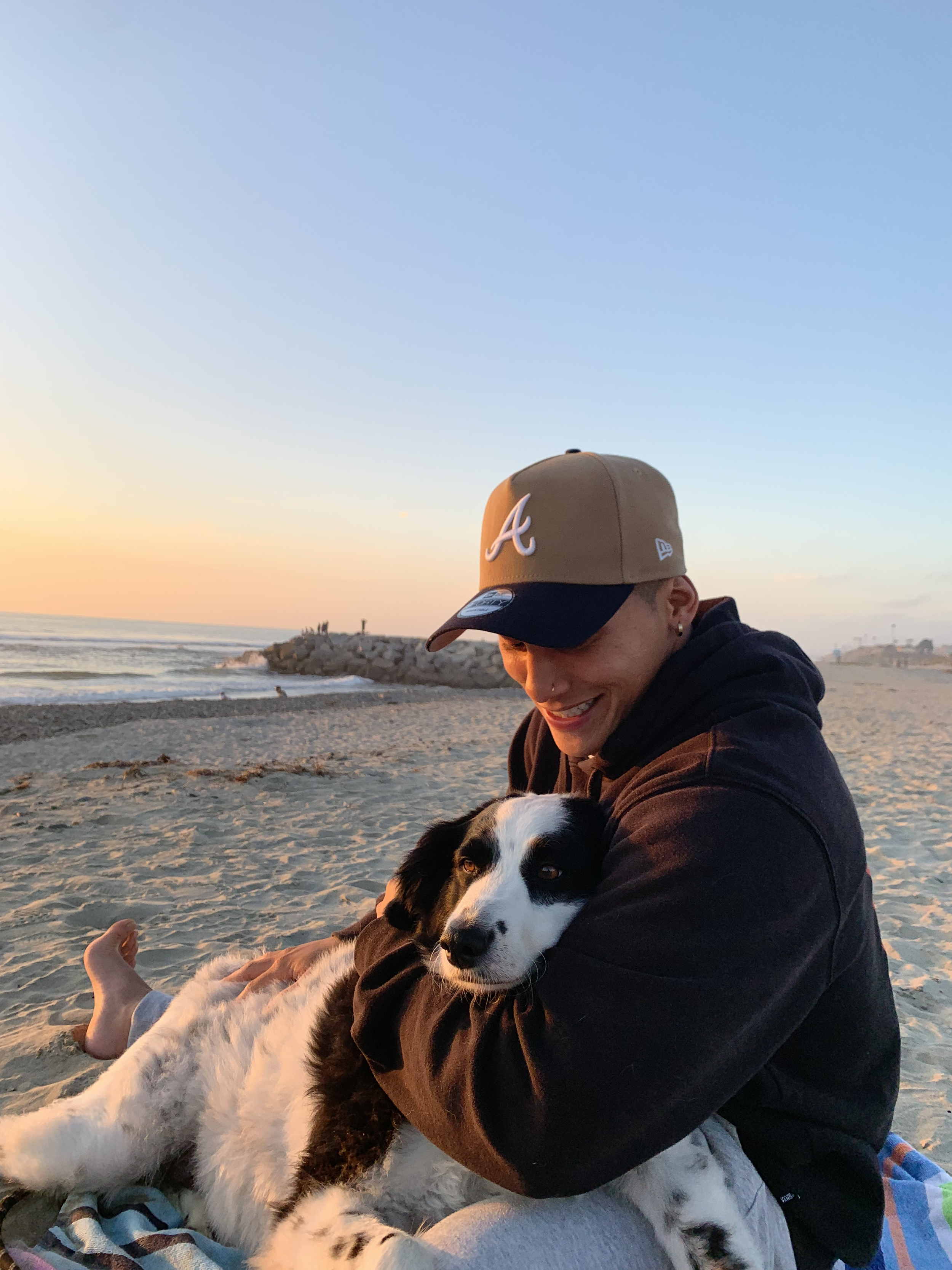 A young man with a cap and hoodie smiling while hugging a black and white dog on a beach during sunset.