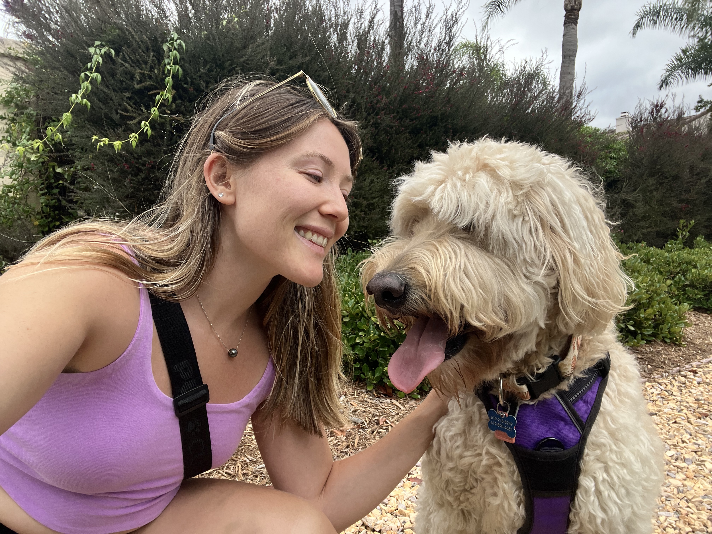 A woman smiling and crouching next to a large, fluffy, light-colored dog with curly fur, wearing a purple harness, outdoors with greenery and trees in the background.